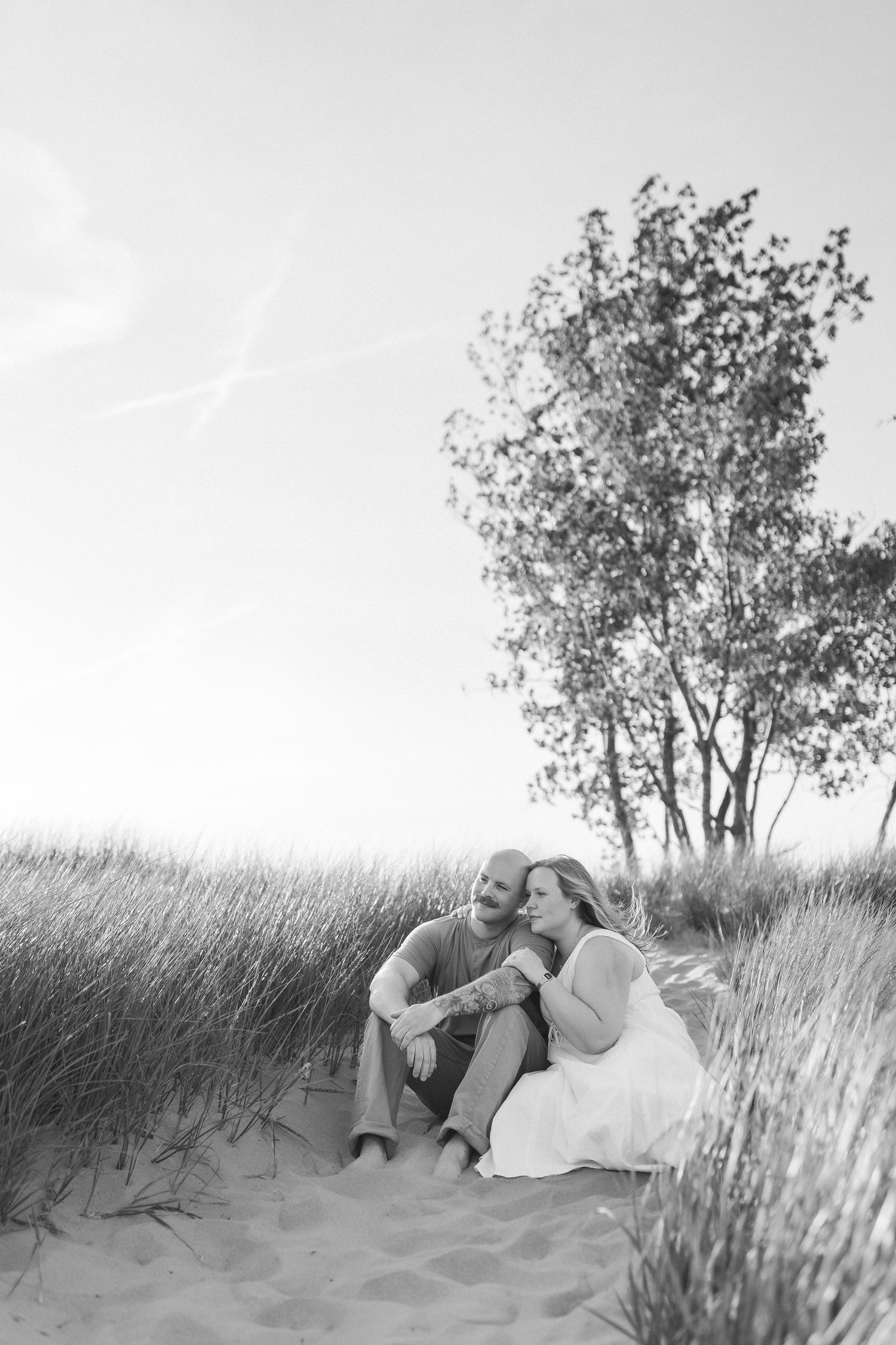 A romantic black and white photo of a couple sitting together on a sandy dune with tall grass and a tree in the background.