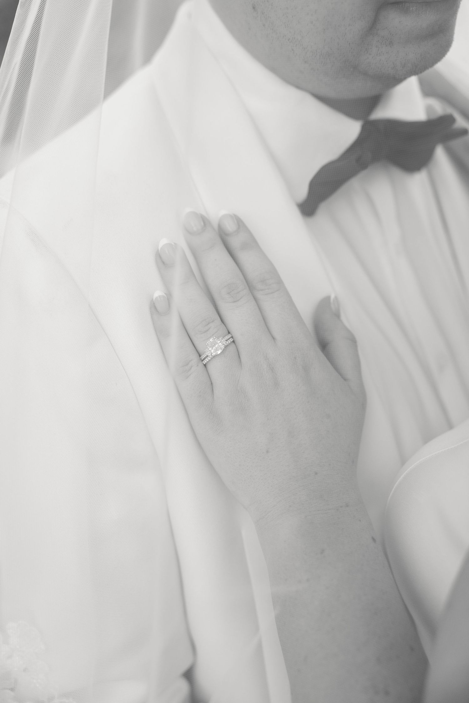 Close-up of bride's hand with diamond engagement ring resting on groom's white tuxedo shirt.