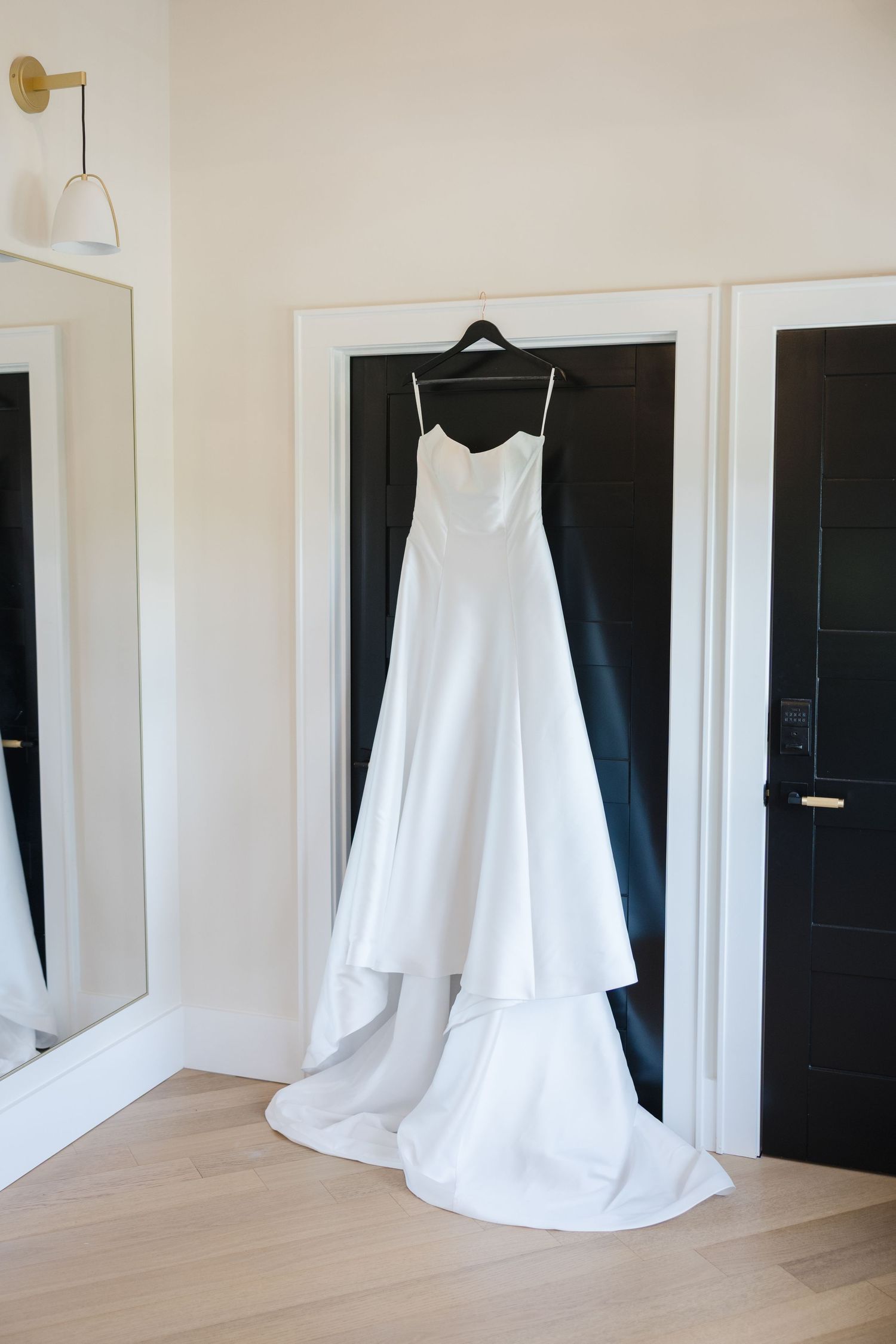 Elegant white satin A-line wedding dress with train hanging on black door in bright bridal suite.
