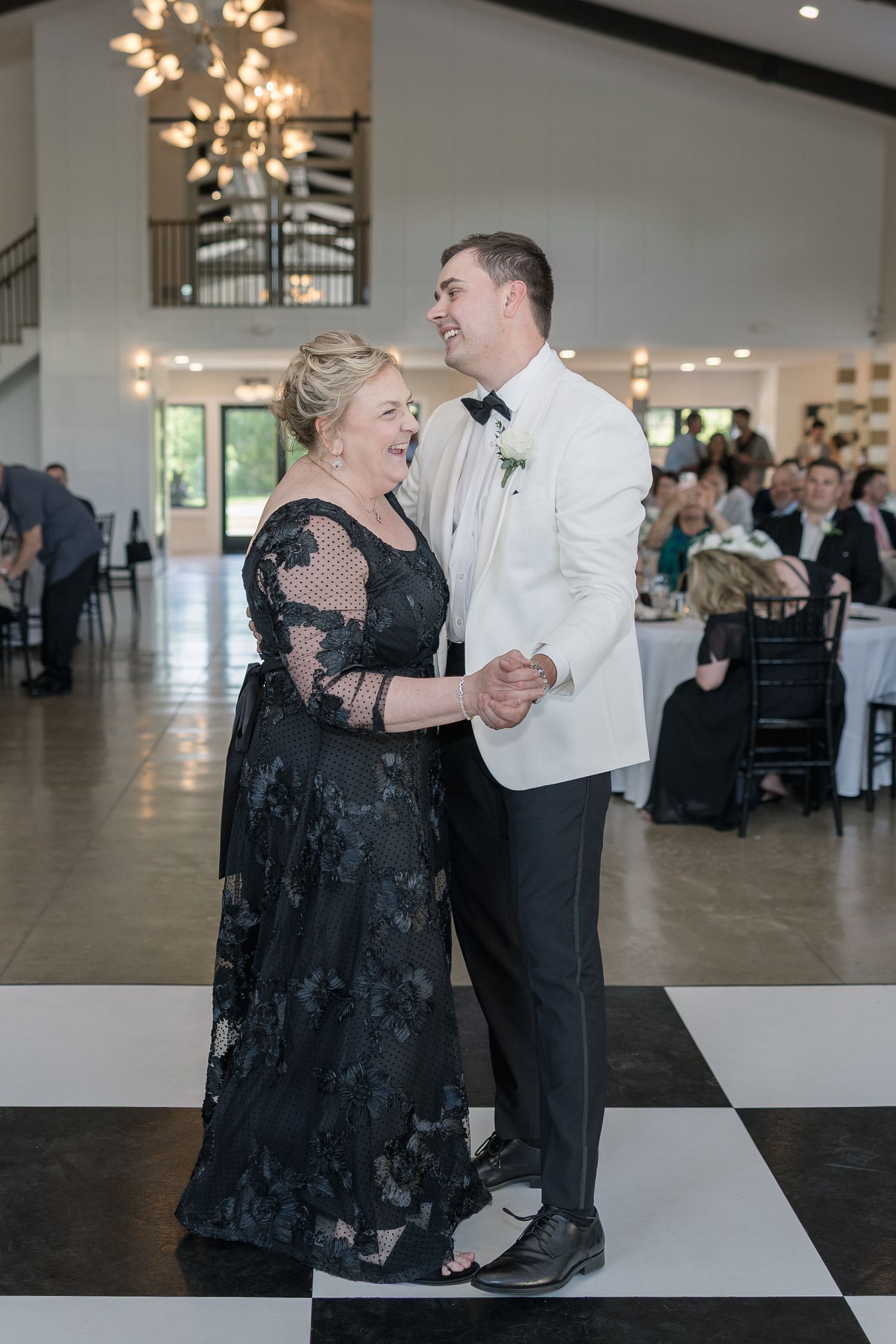 Groom in white tuxedo shares mother-son dance on checkered floor at elegant wedding reception.