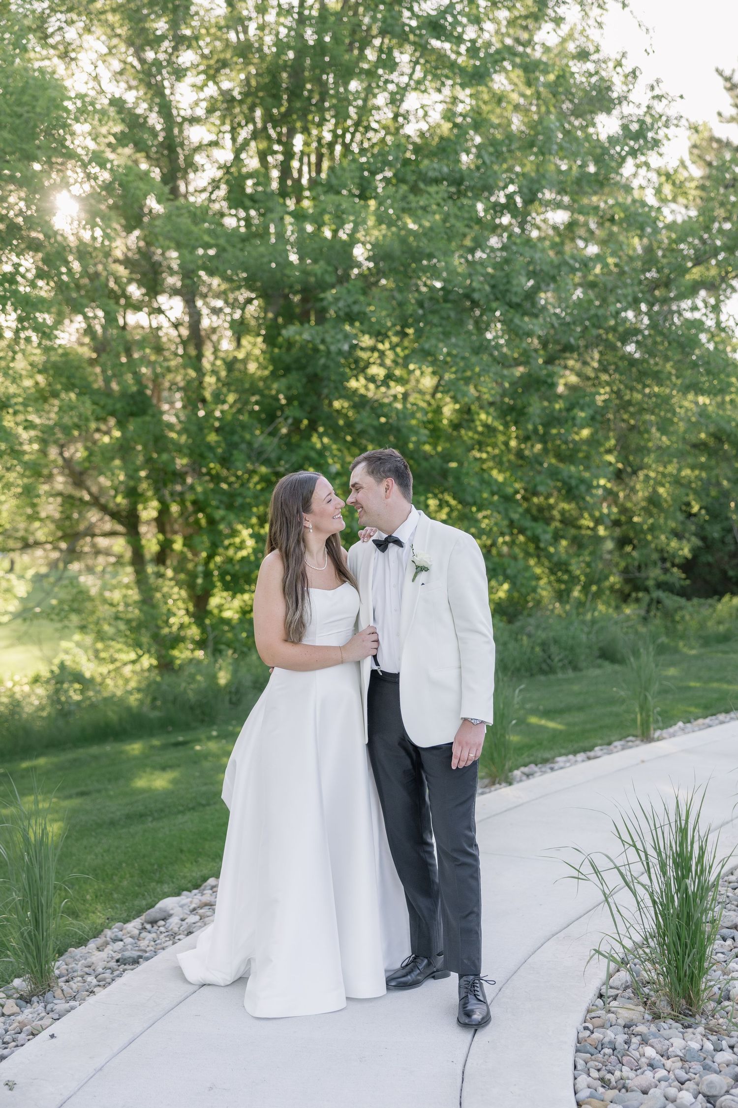 Bride and groom sharing a loving glance while walking on sunlit garden path surrounded by lush green trees.