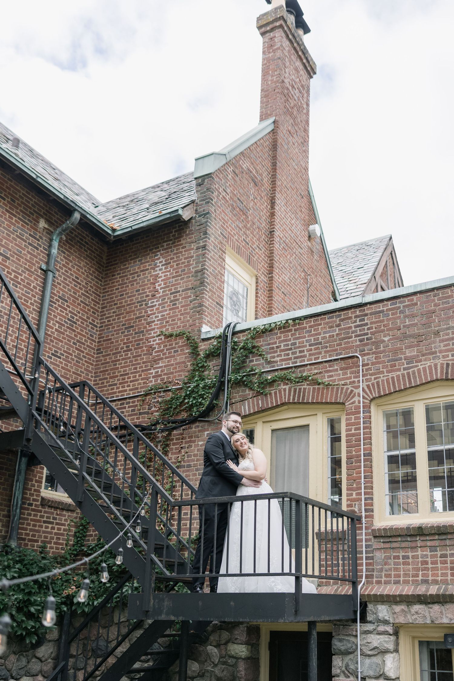 Couple embraces on black metal staircase landing against brick mansion with tall chimney and stone foundation.