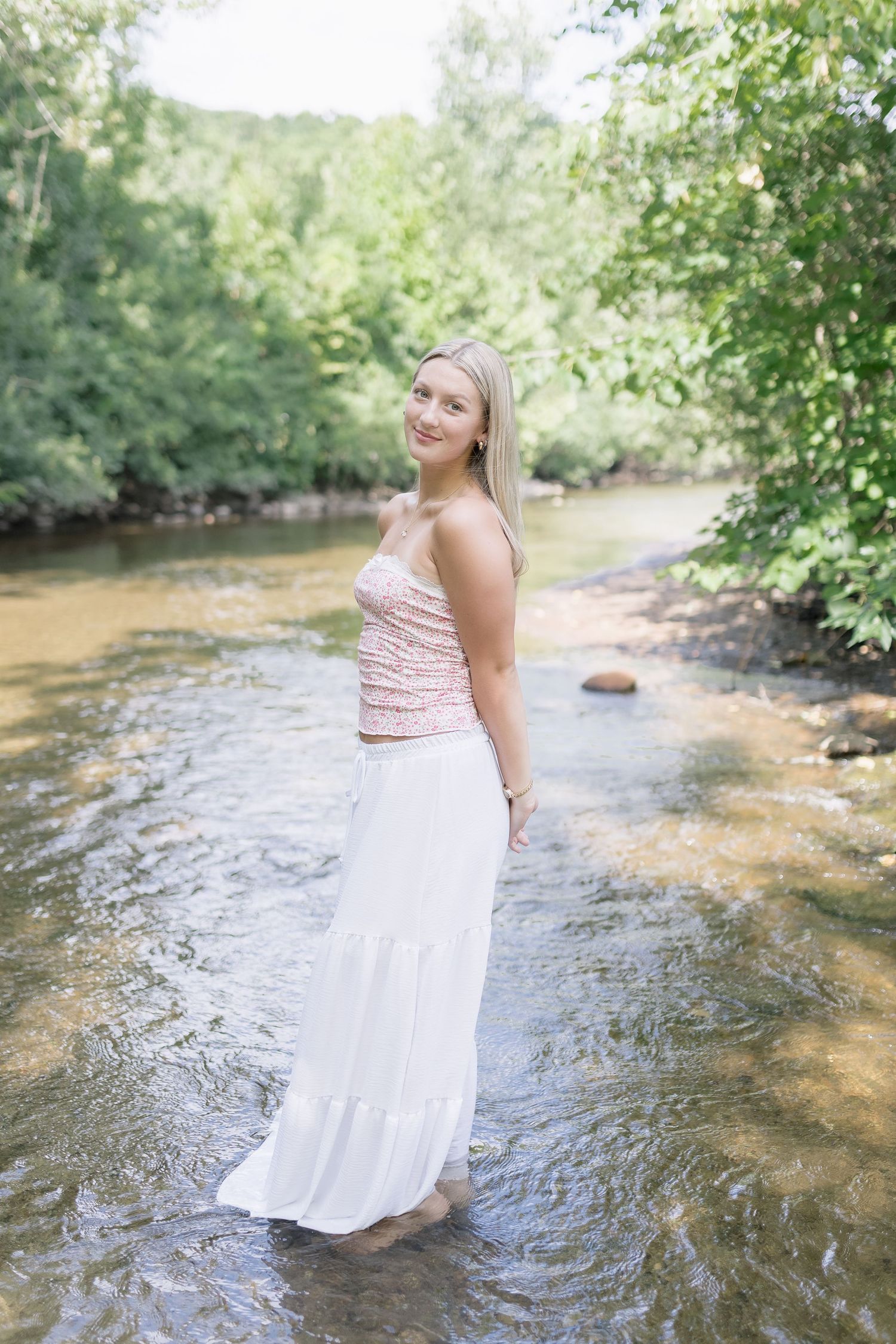 A person in a striped top and white skirt sits gracefully in a flowing creek surrounded by lush greenery.