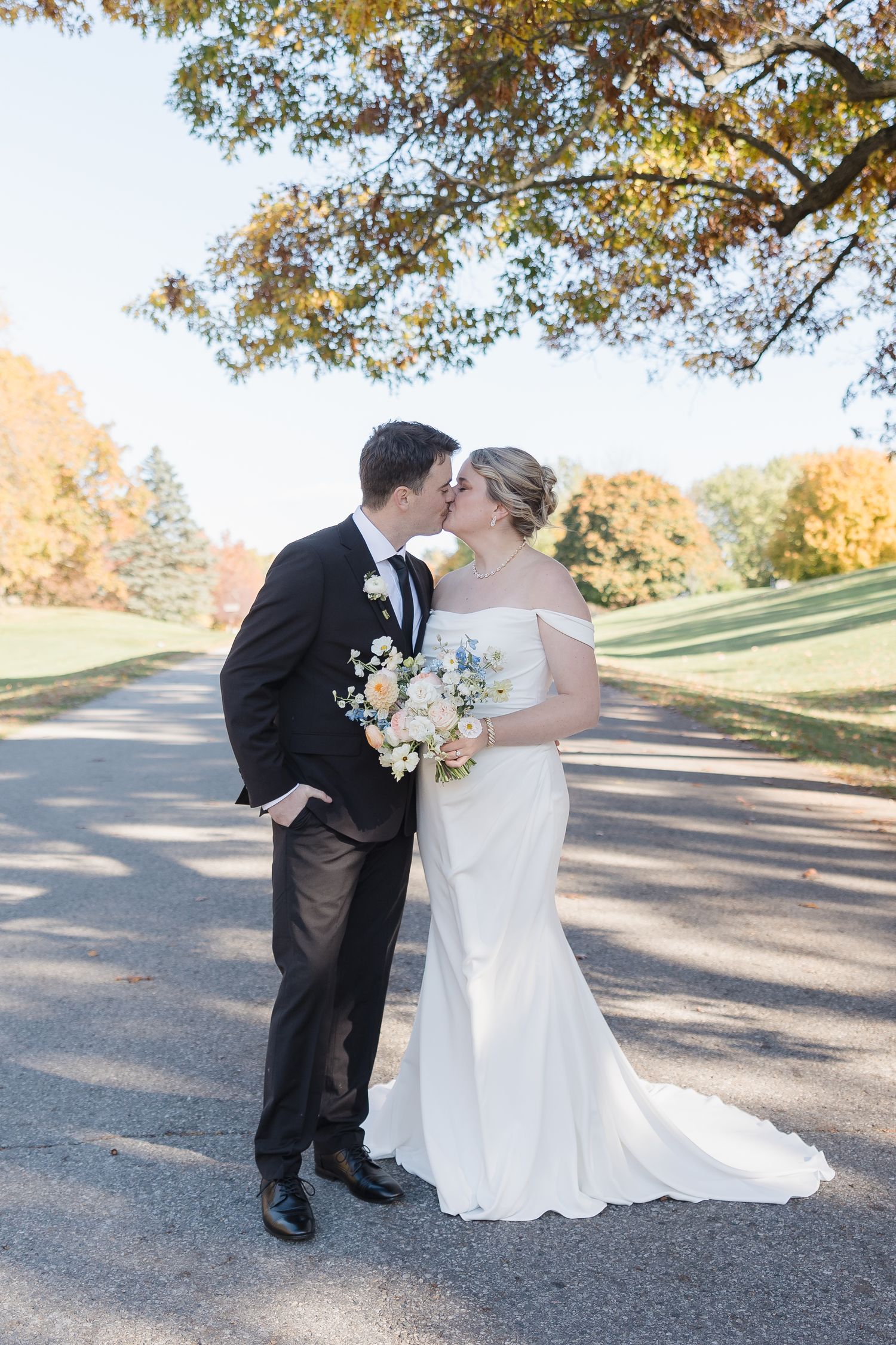 Romantic wedding couple embraces on tree-lined path with beautiful fall foliage.