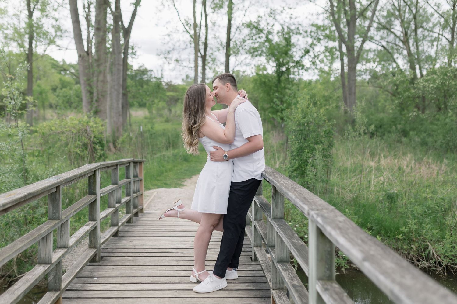 A couple embraces while walking along a scenic wooden boardwalk path through lush green forest surroundings.