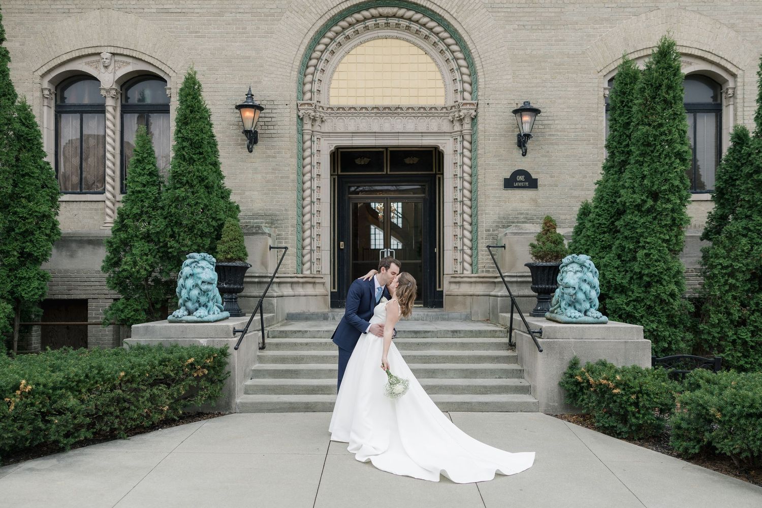 Bride and groom share a romantic kiss outside an elegant stone building with blue lion statues and lush greenery.