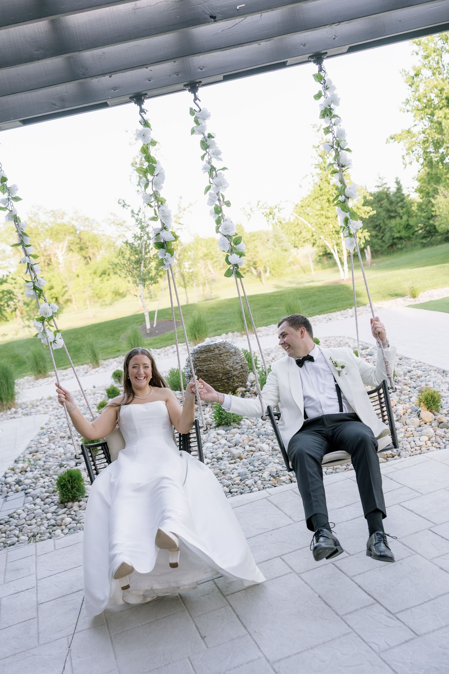 Bride and groom laugh together while sitting on decorated swings under a pergola on their wedding day.