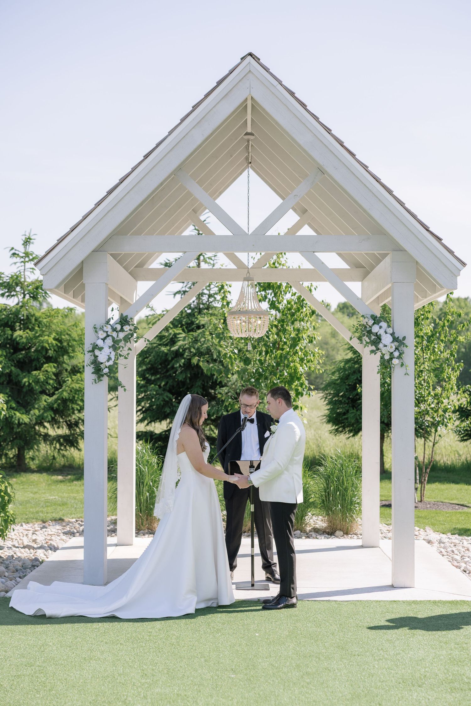 Bride and groom exchange vows under a white gazebo adorned with flowers during an outdoor wedding ceremony.
