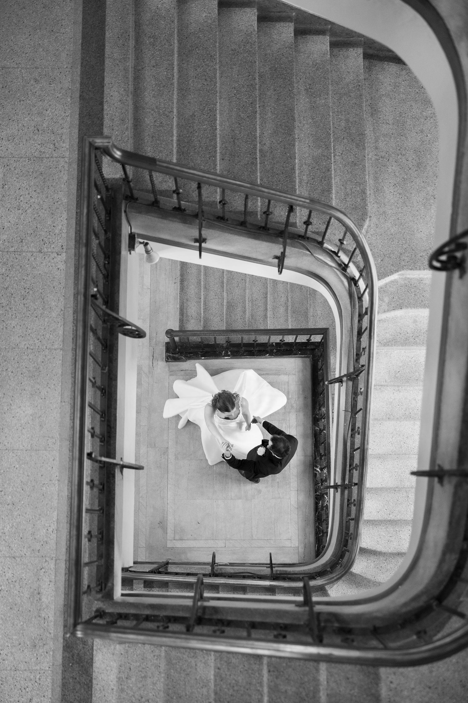 Aerial black and white photo of a bride and groom embracing at the base of an elegant spiral staircase.
