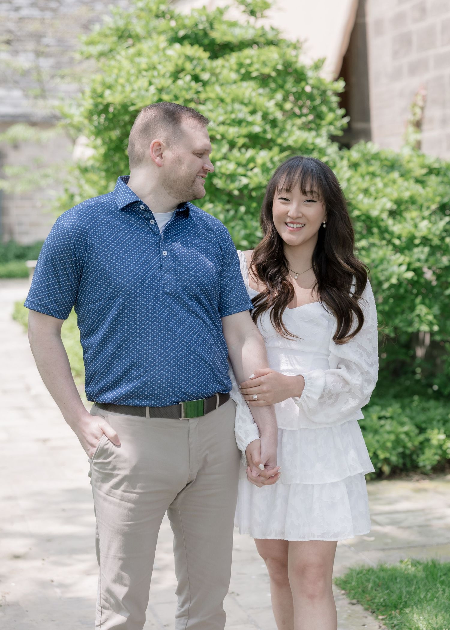 A couple shares a romantic moment together on a sunny day outdoors near a stone wall and greenery.