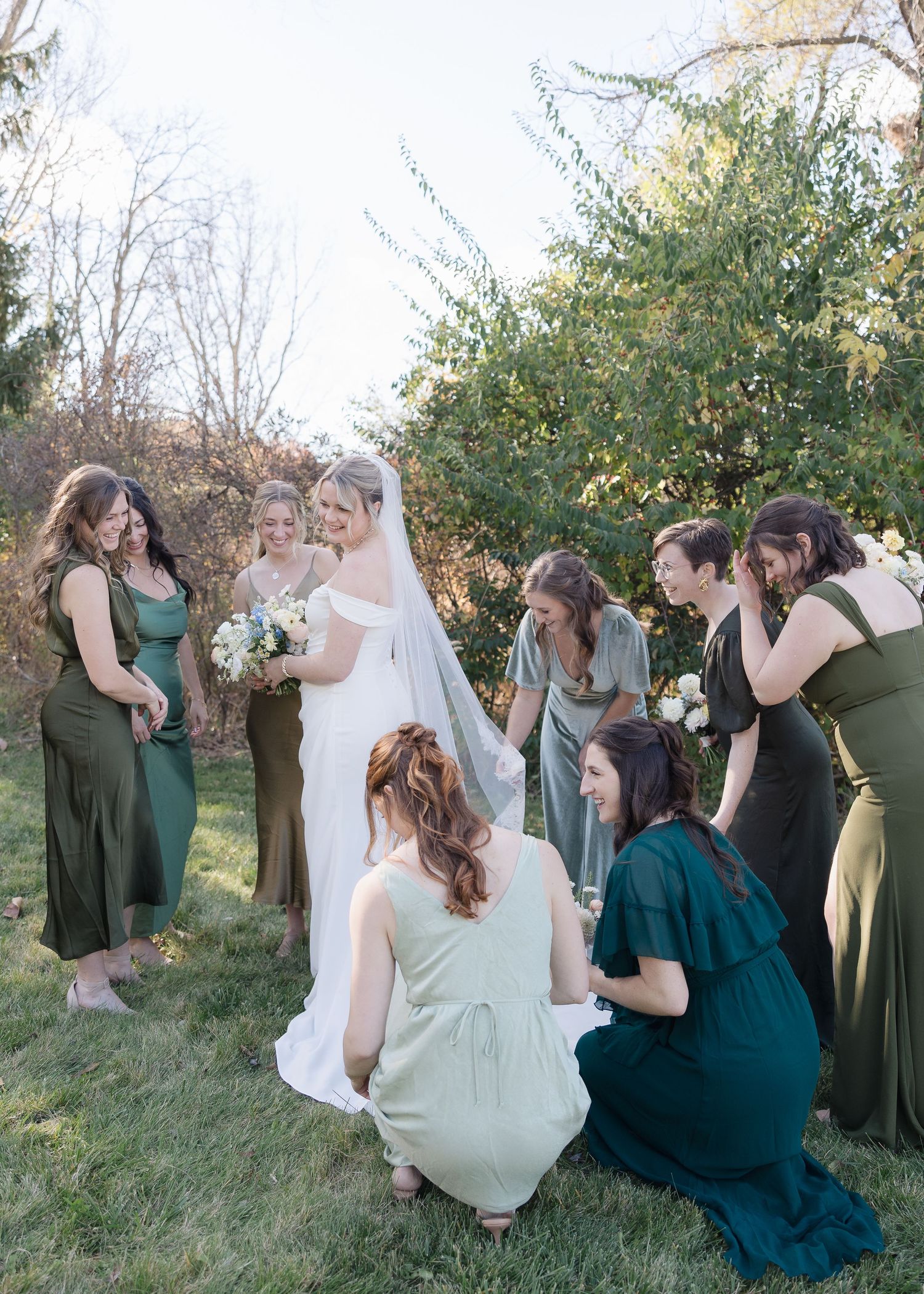 Wedding party gathers around bride in white dress for candid outdoor photo moment.