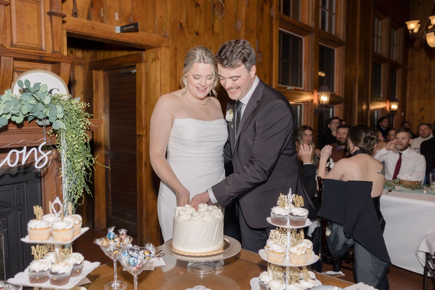 A couple cuts their white wedding cake together at a rustic wooden venue with dessert displays.