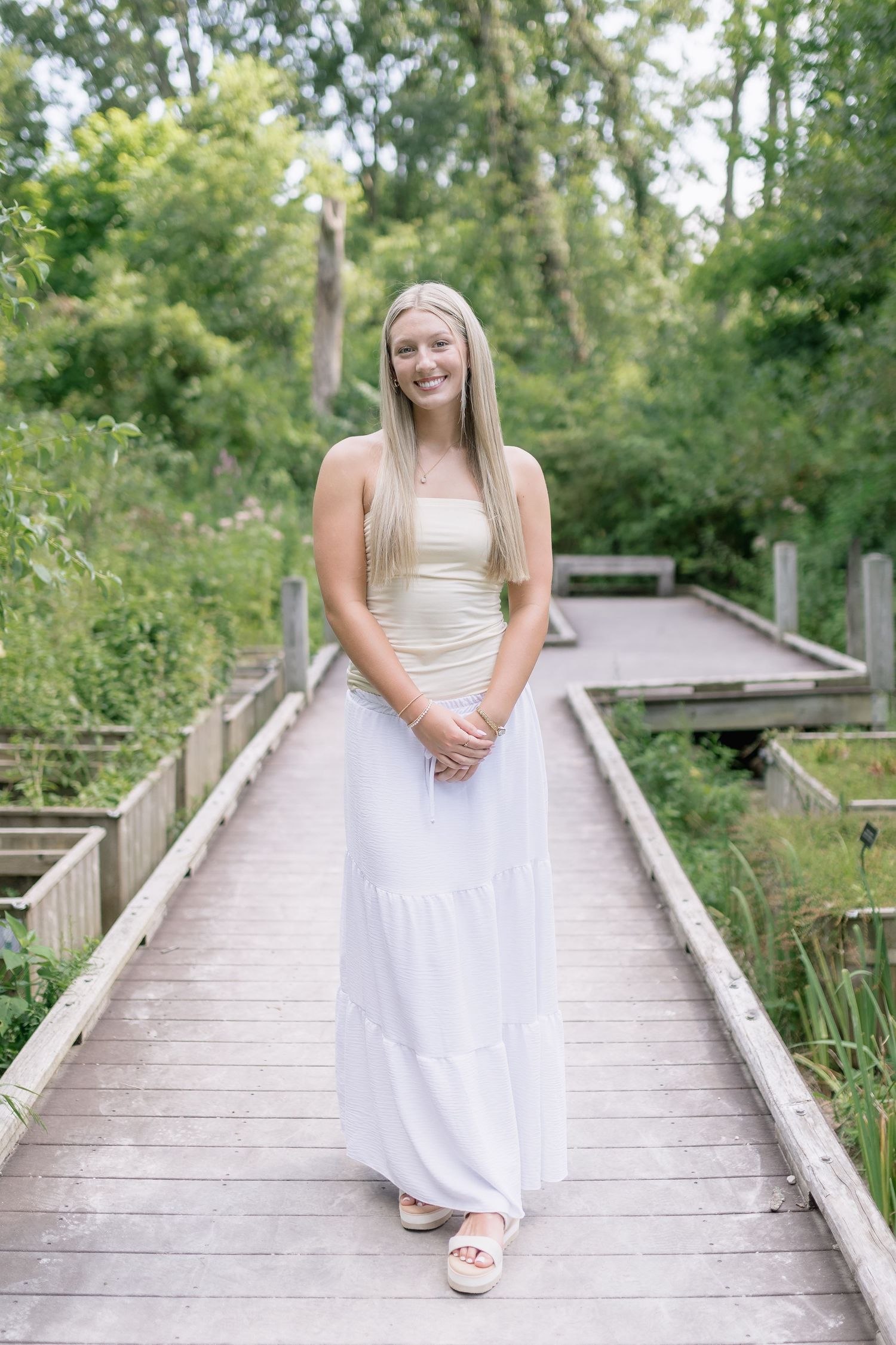 A person in a white dress sits on a rustic wooden boardwalk surrounded by lush summer greenery.