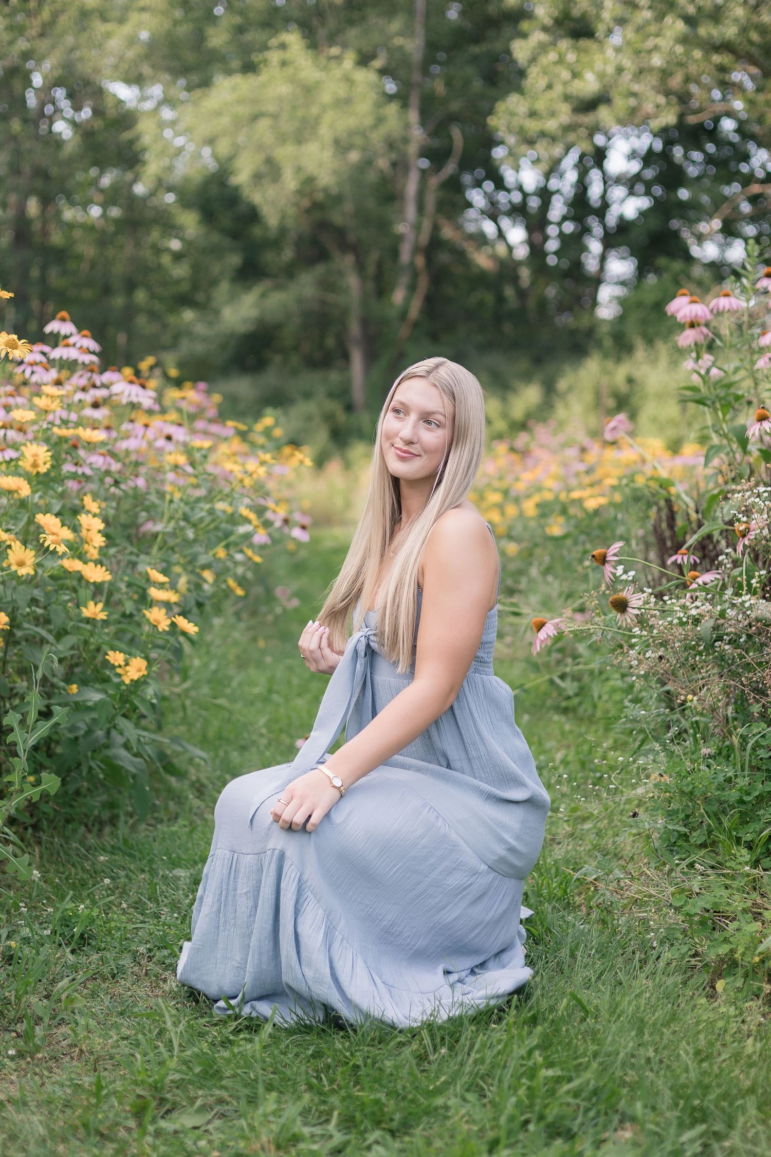 A person in a flowing blue dress sits gracefully among colorful wildflowers in a lush summer garden setting.