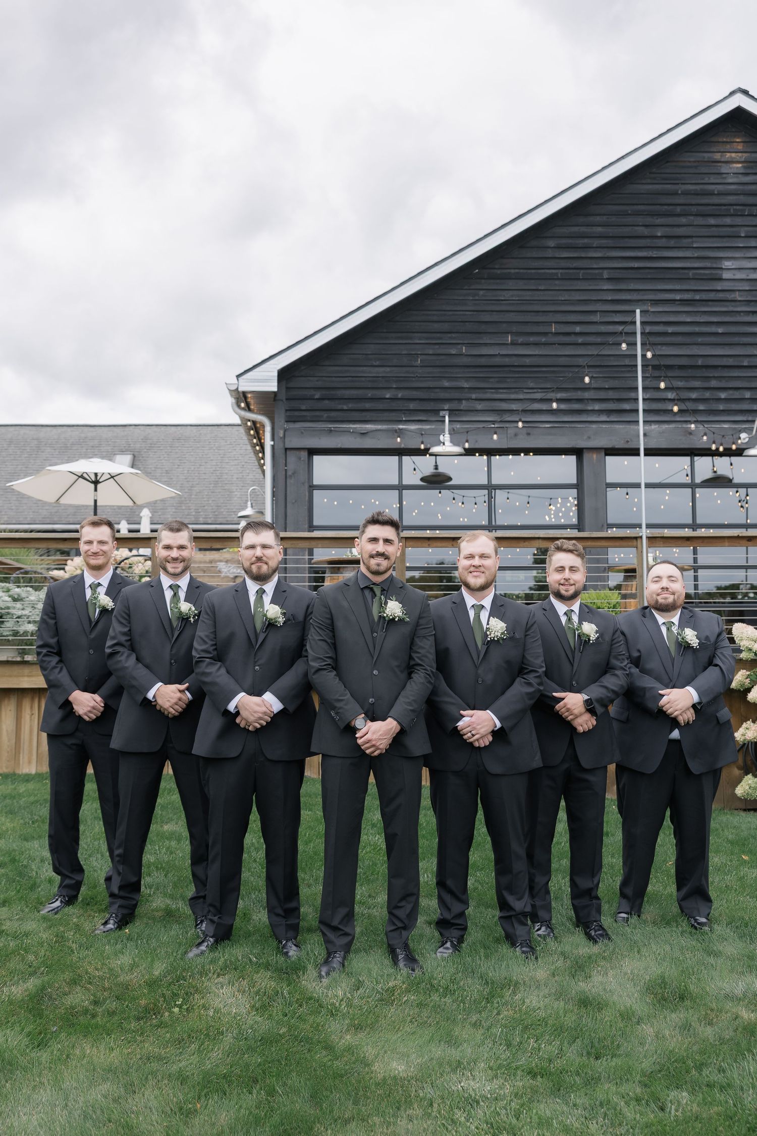 A wedding party of groomsmen in black suits stand in a row against a dark barn exterior on a cloudy day.