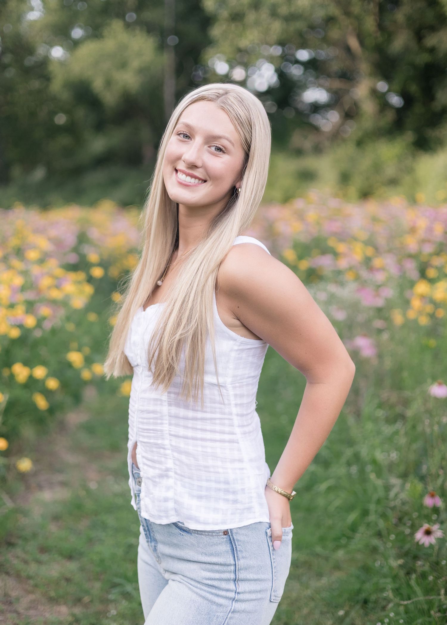 A person in a white sleeveless top and light jeans poses in a field of colorful wildflowers on a sunny summer day.