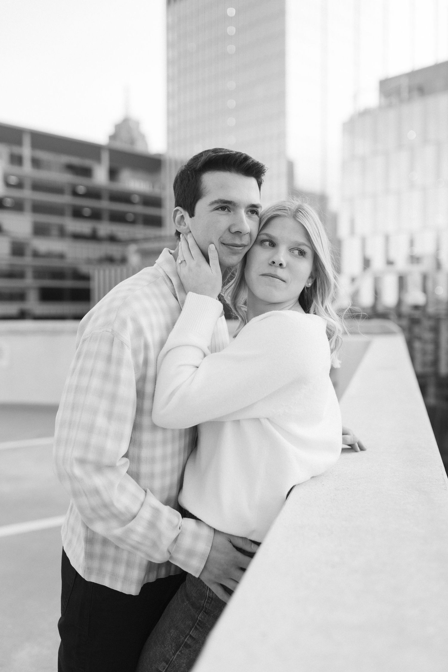 A black and white portrait of a couple embracing with city buildings in the background.