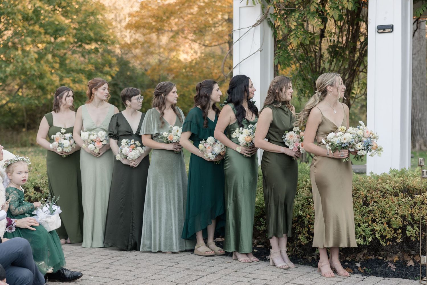 Bridesmaids in mixed green and neutral dresses holding bouquets at an outdoor ceremony.