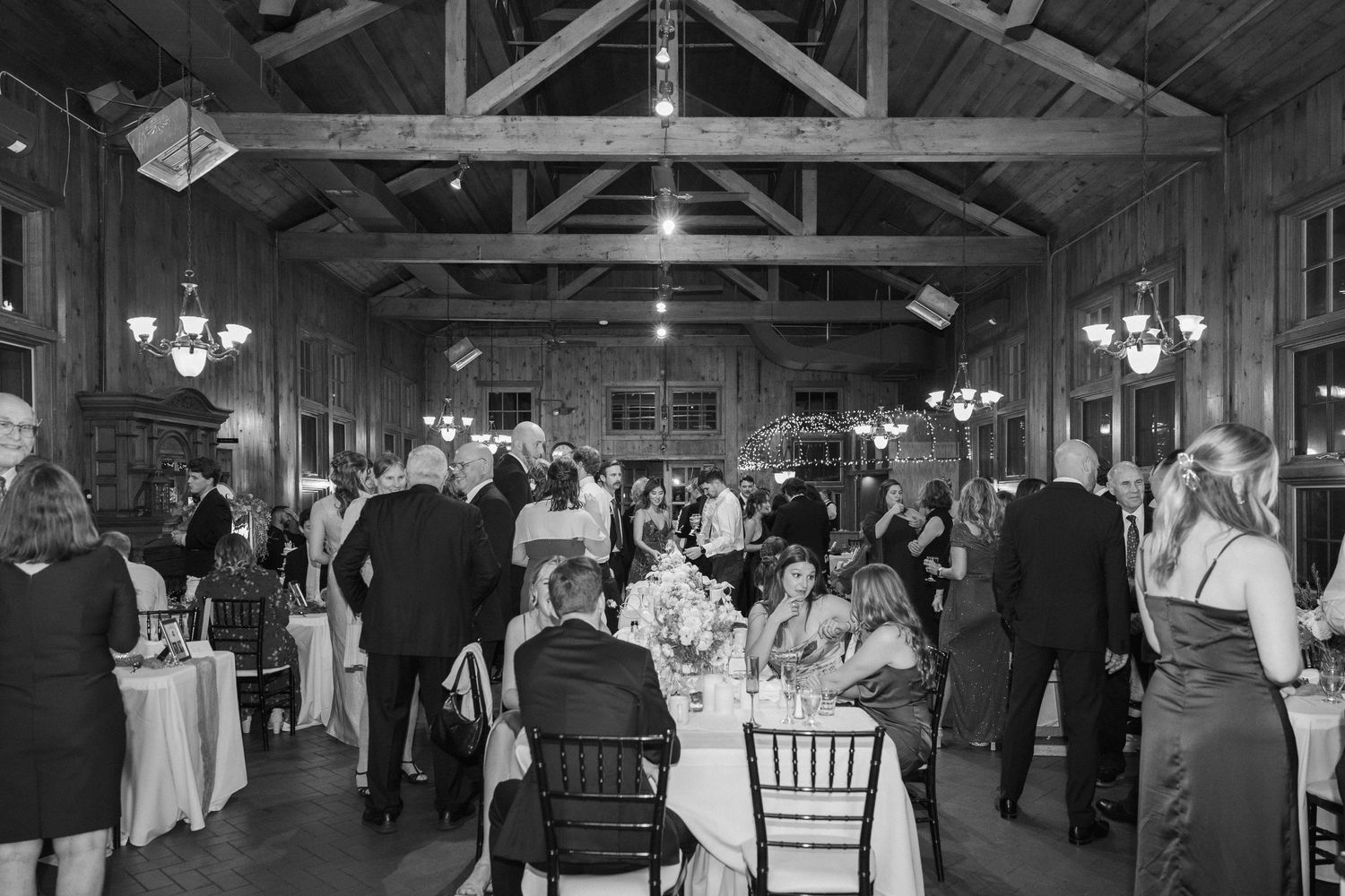 Black and white photo of wedding reception inside rustic barn venue with exposed beams and hanging lights.