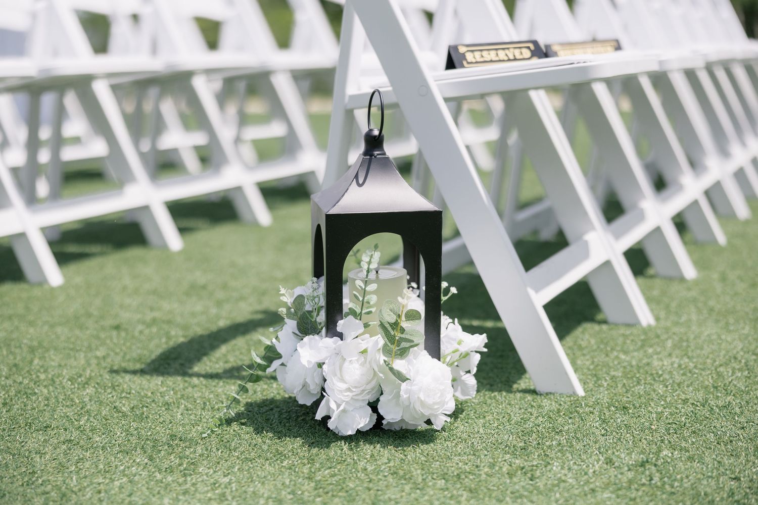 Black lantern with white roses on grass beside white folding chairs at an outdoor wedding ceremony.