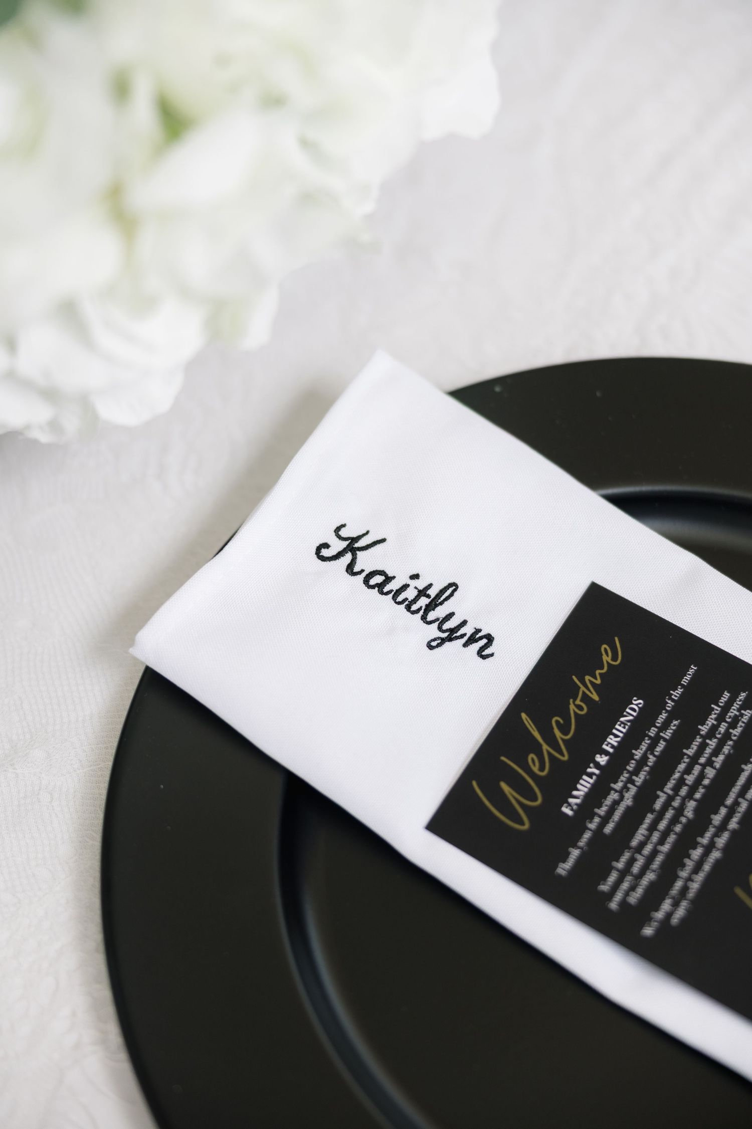 Personalized white napkin with Kaitlyn embroidered in script on black charger plate with welcome card.