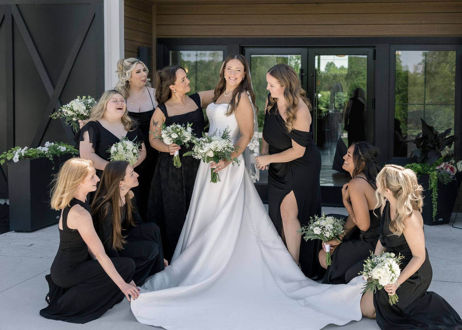 Bride in white gown surrounded by bridesmaids in black dresses holding white bouquets outside a modern venue.