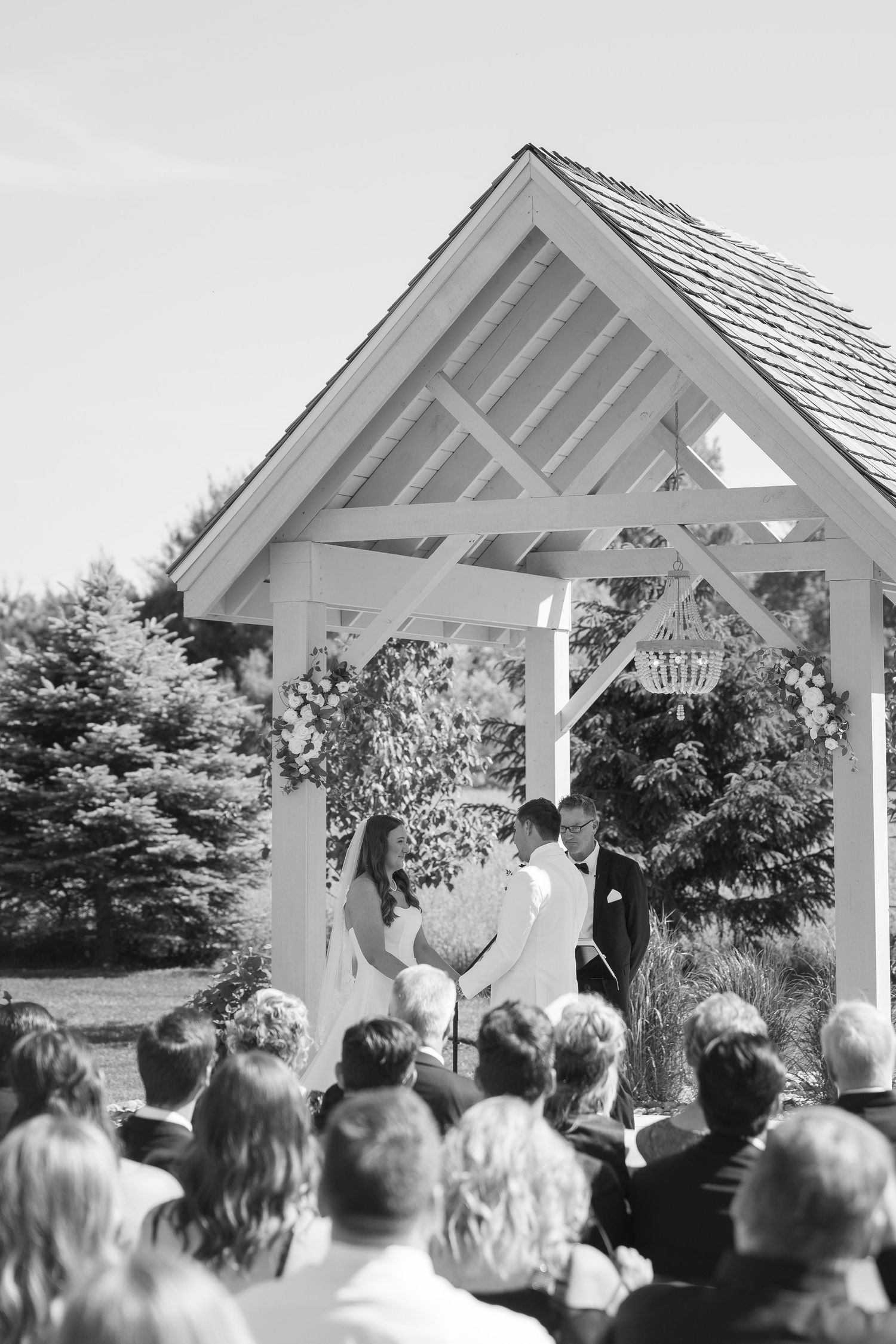 Black and white outdoor wedding ceremony under a white wooden gazebo with guests seated watching the couple exchange vows.
