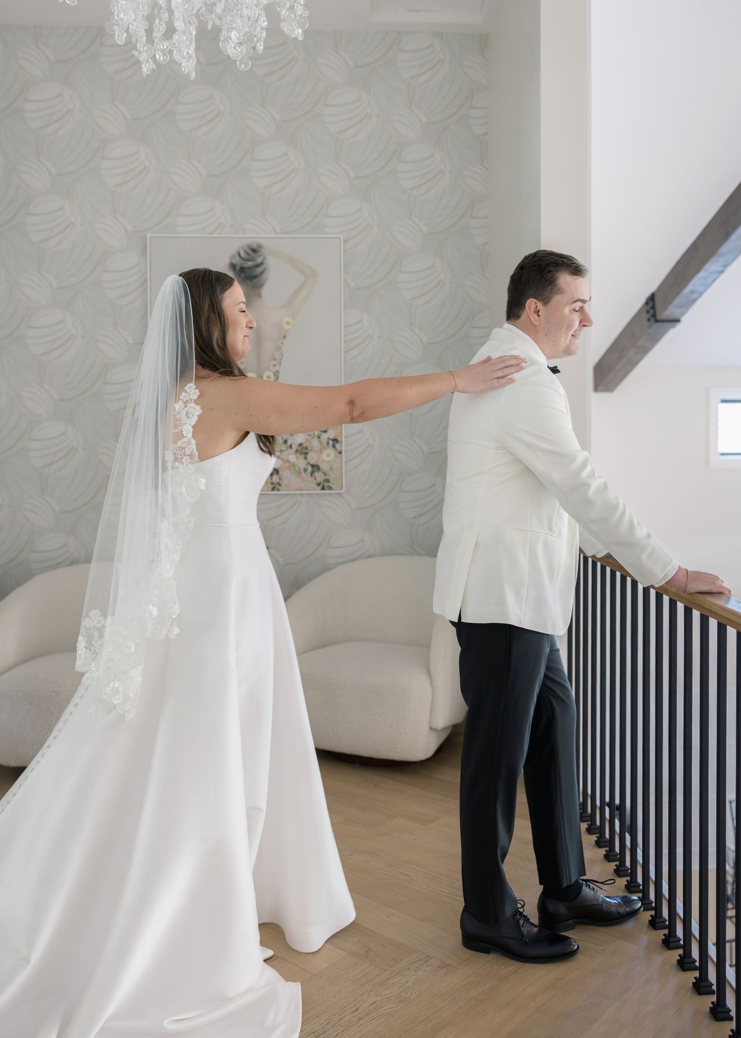 Bride in white gown and veil taps groom's shoulder for first look moment on staircase.