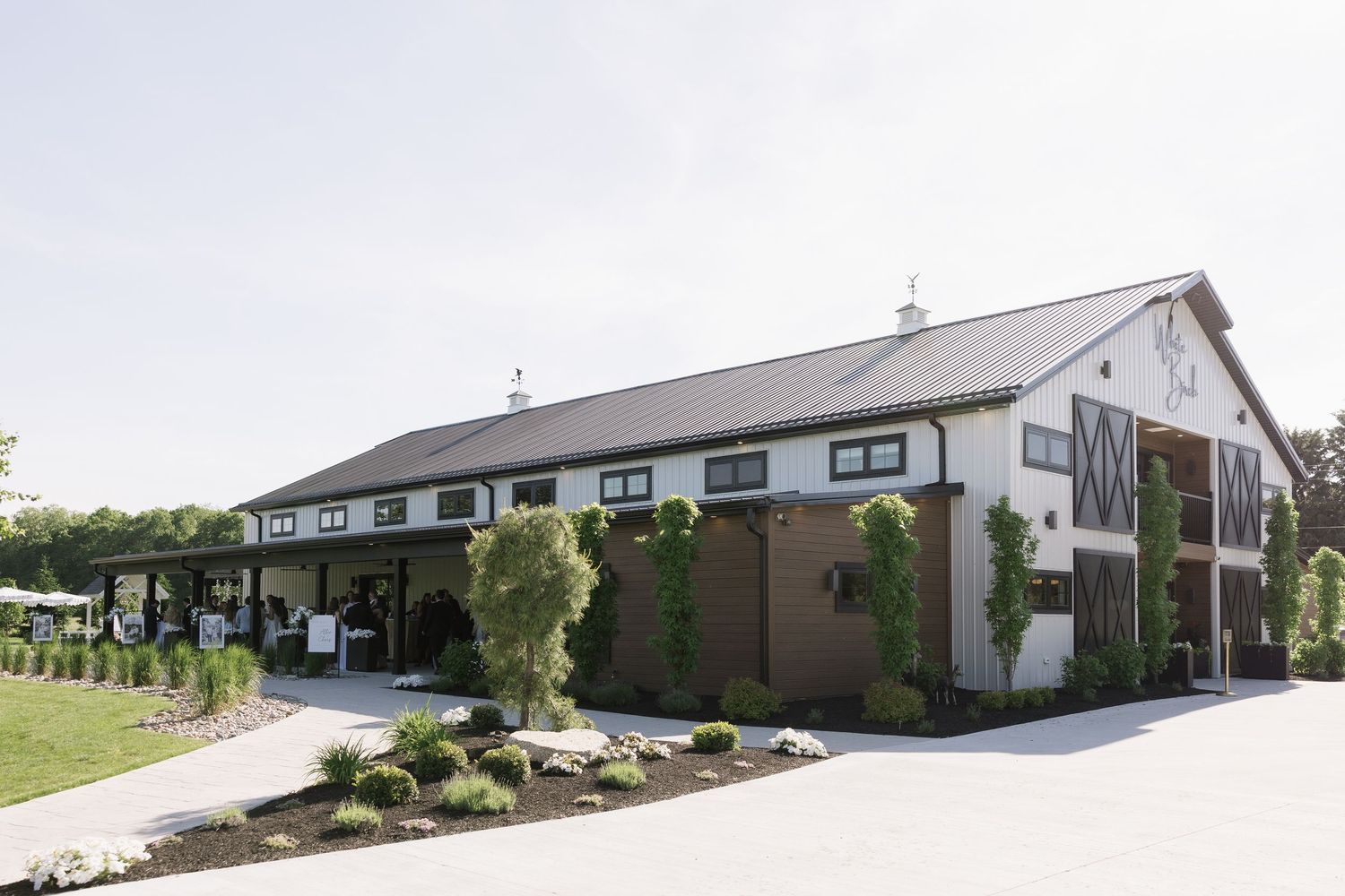 A modern white barn wedding venue with dark trim, lush greenery, and manicured landscaping.