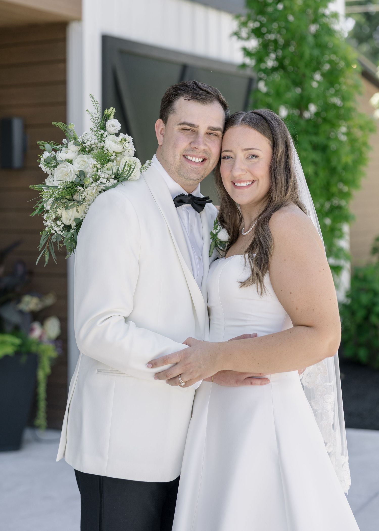 Bride and groom pose together outdoors, she in a white gown with veil, he in a white tuxedo jacket with bow tie.