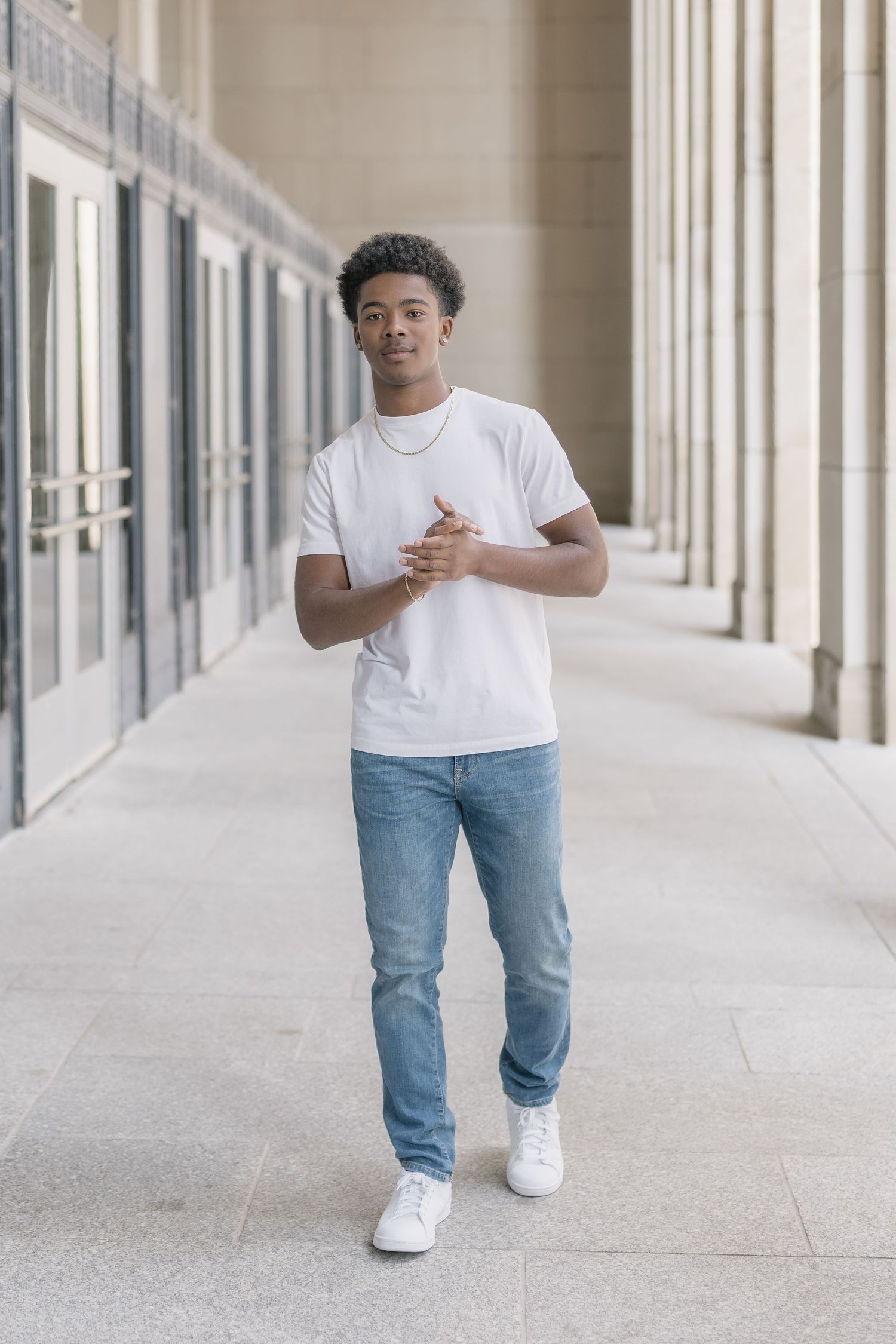 Person in white t-shirt and jeans walking through a bright hallway with columns and white walls.