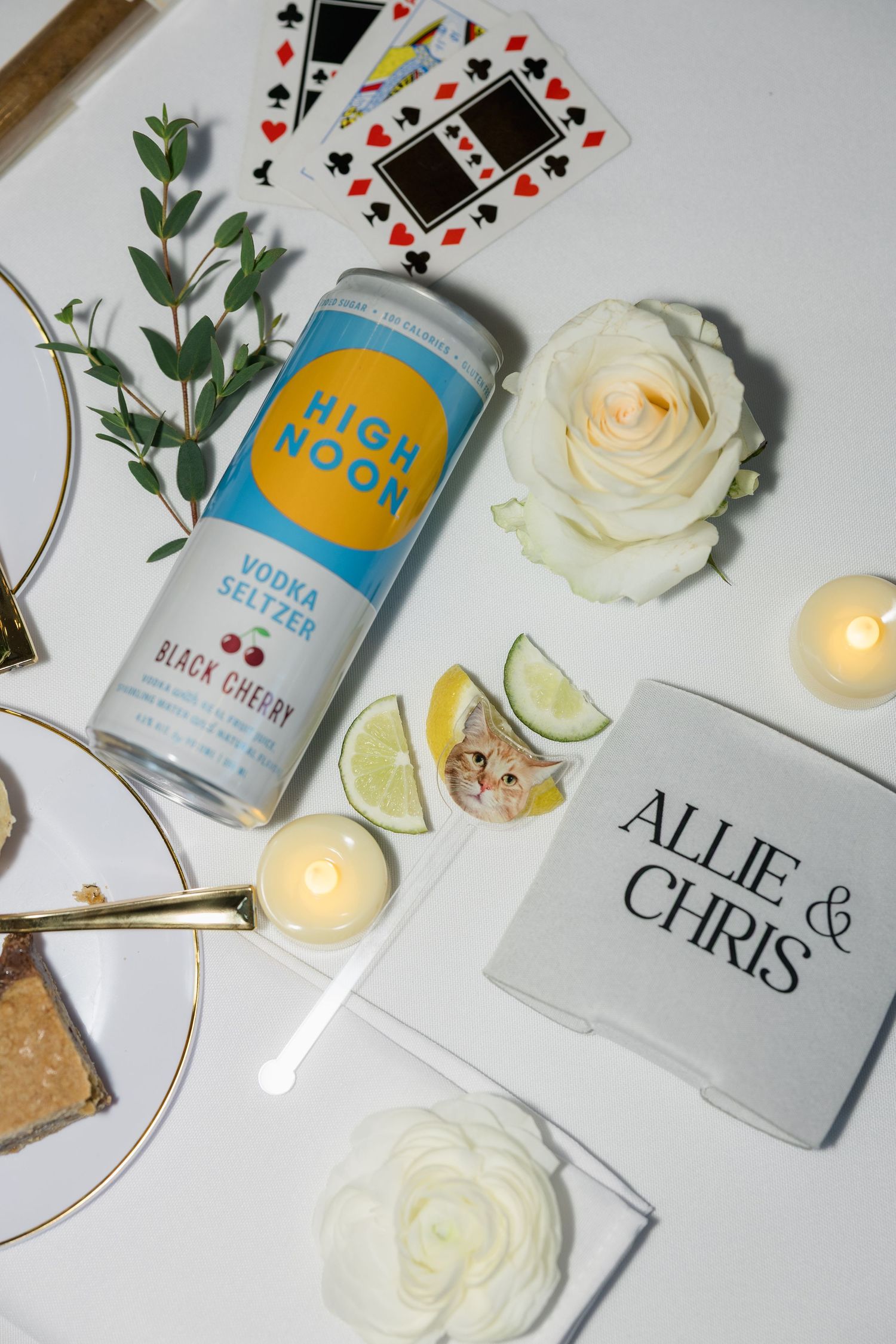Flat lay of wedding details featuring High Noon Black Cherry seltzer, playing cards, white roses, and Allie & Chris signage.