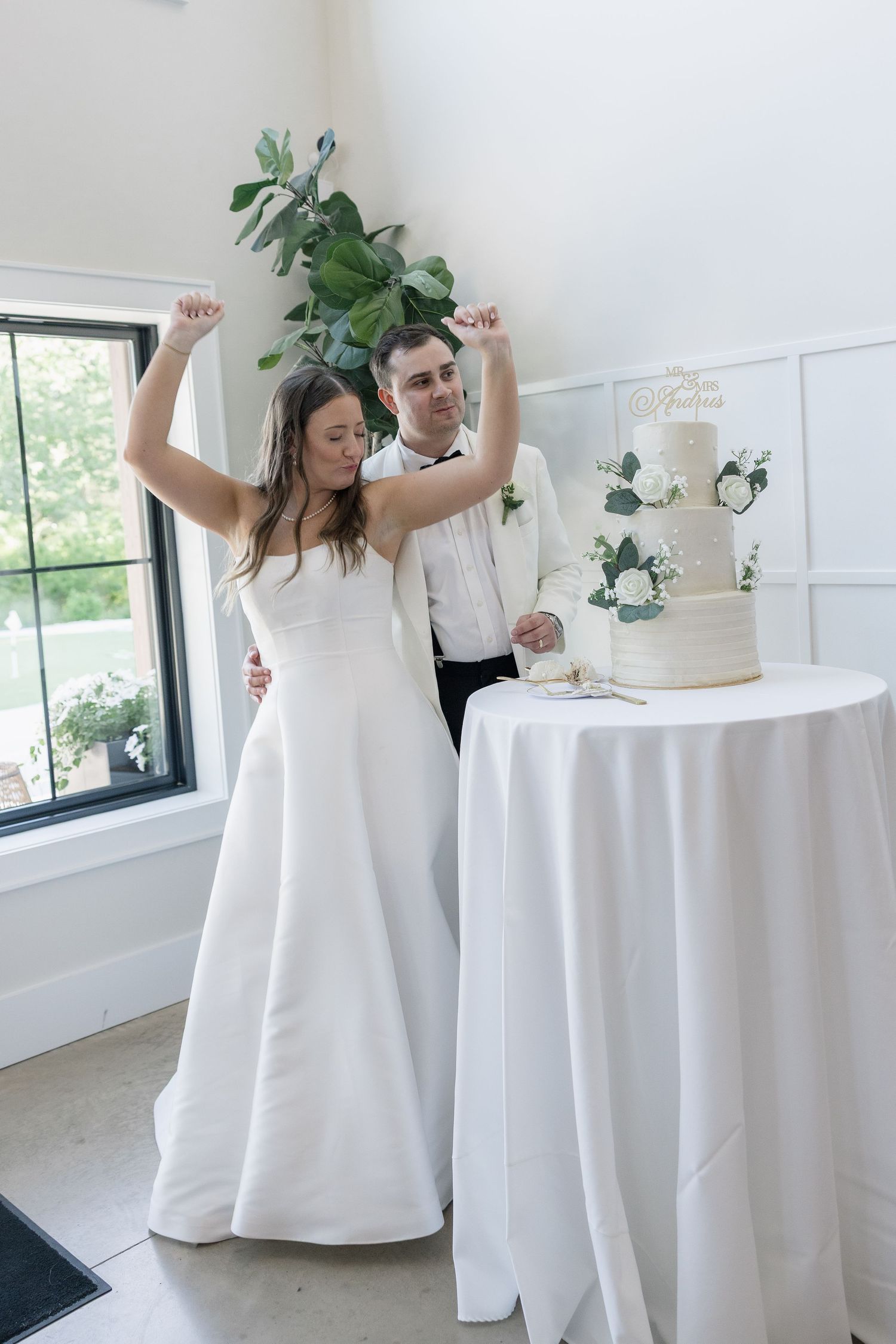 Bride and groom celebrate cutting their white floral wedding cake, raising fists in joy at elegant indoor venue.