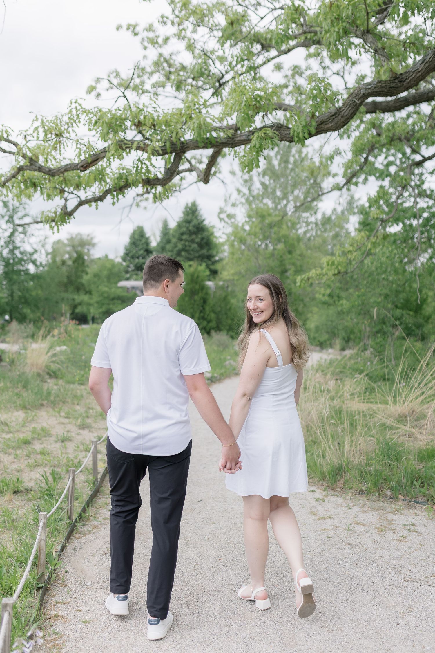 A couple walks hand in hand along a winding gravel path surrounded by lush summer greenery.