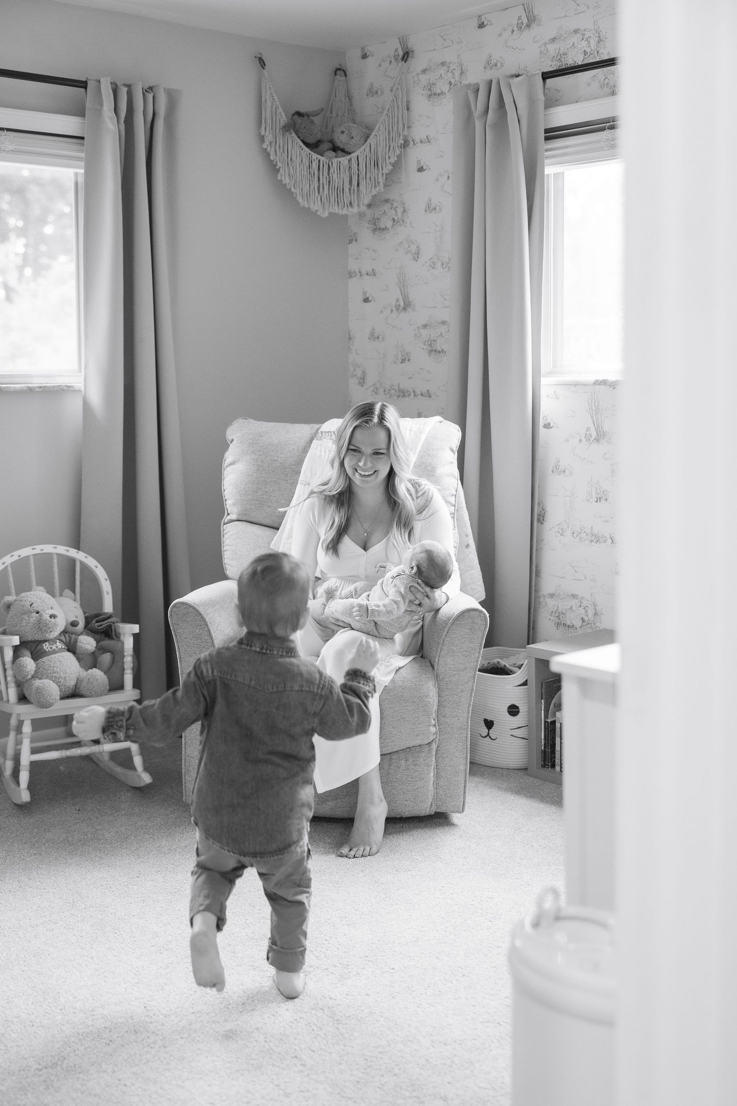 A toddler in gray clothing walks across a bright nursery room with white furniture.