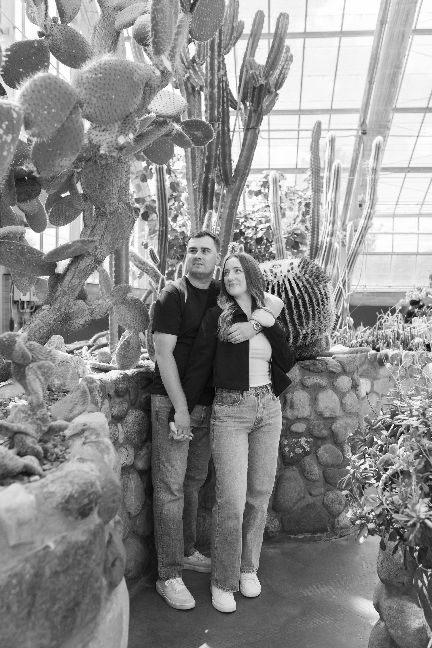 A couple poses together in front of tall cacti and succulents inside a greenhouse conservatory.