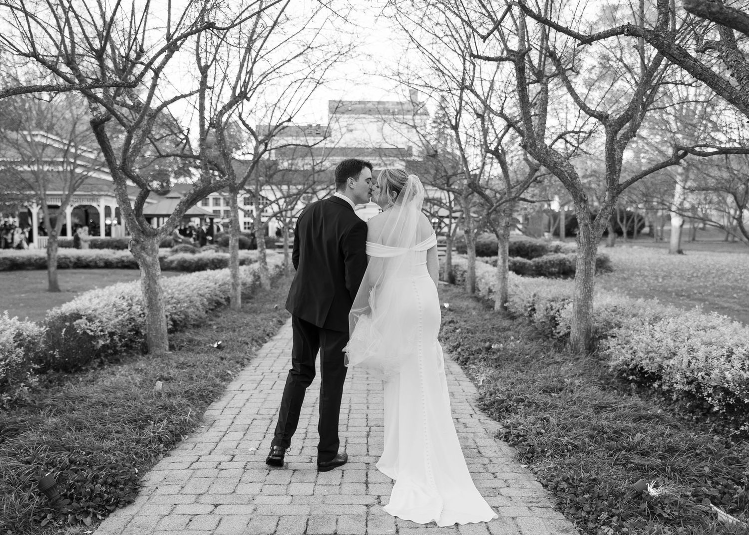 Back view of newlyweds walking down tree-lined brick pathway in garden setting.