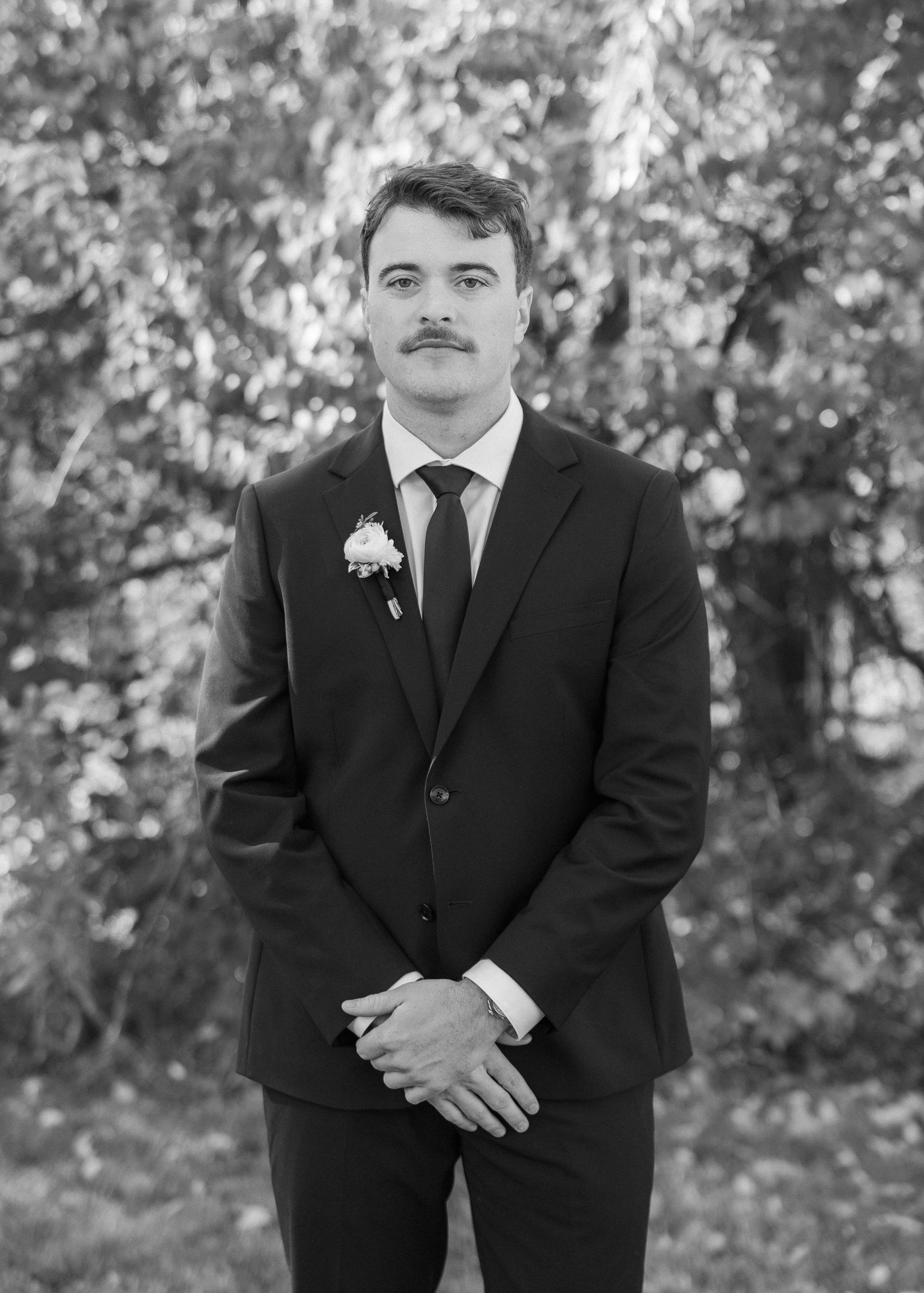 Black and white portrait of a person in a formal suit with a boutonniere standing outdoors against a blurred nature background.