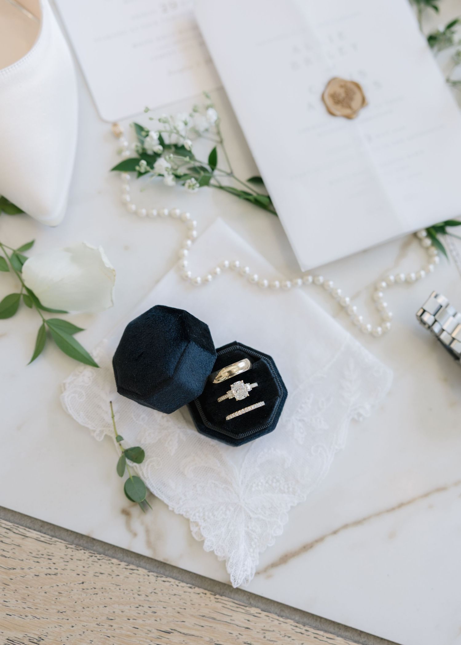 Navy velvet octagonal ring box with gold wedding rings on lace, surrounded by pearl necklace and greenery.