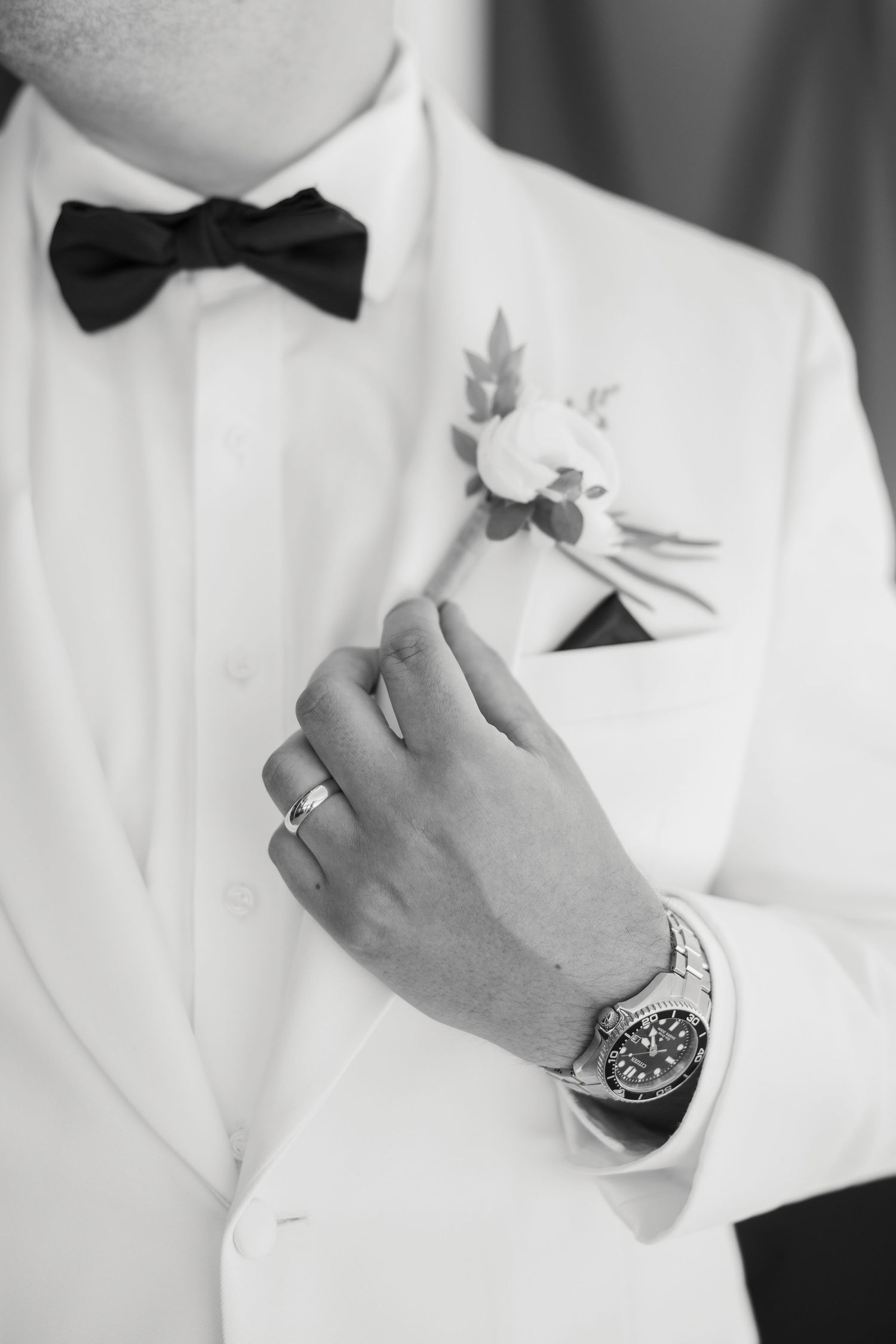 Black and white close-up of groom in white tuxedo adjusting boutonniere, wearing watch and wedding ring.