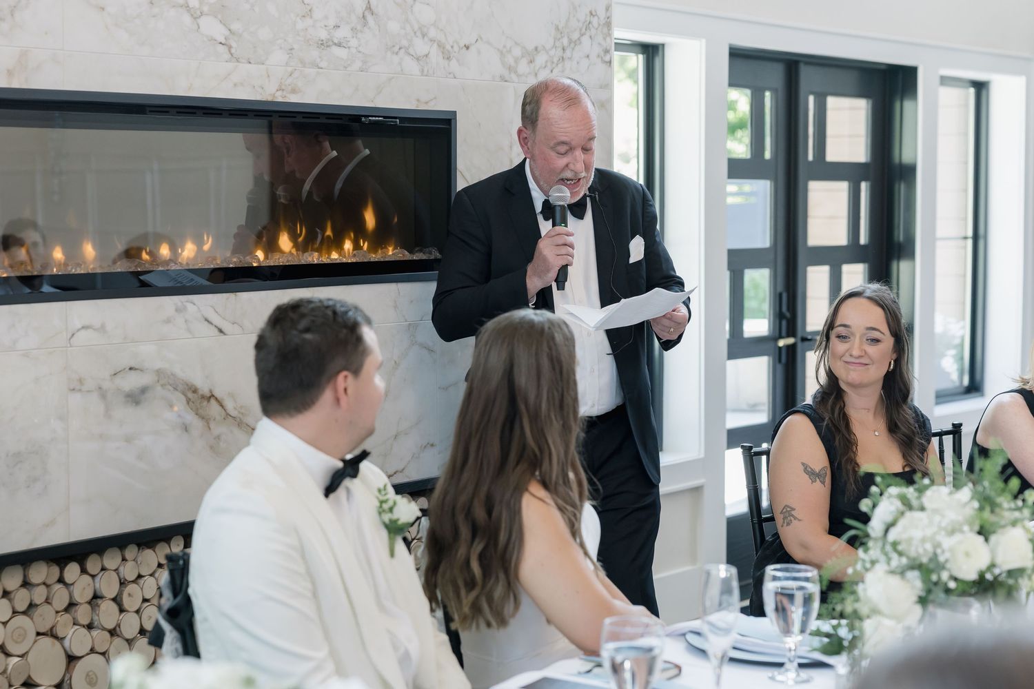 Father of the bride gives a heartfelt speech at an elegant wedding reception with a marble fireplace backdrop.