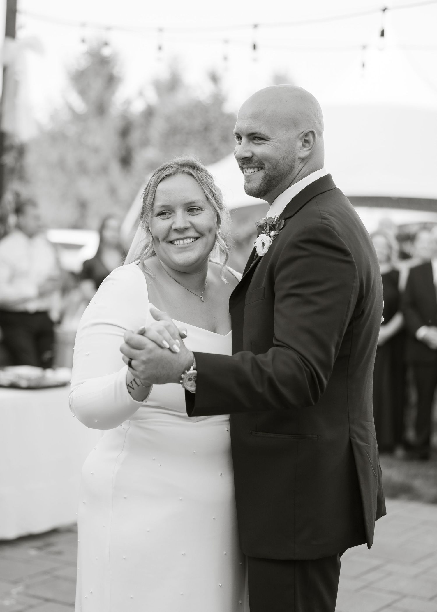Bride and groom share first dance outdoors, smiling joyfully, in elegant wedding attire, black and white photo.