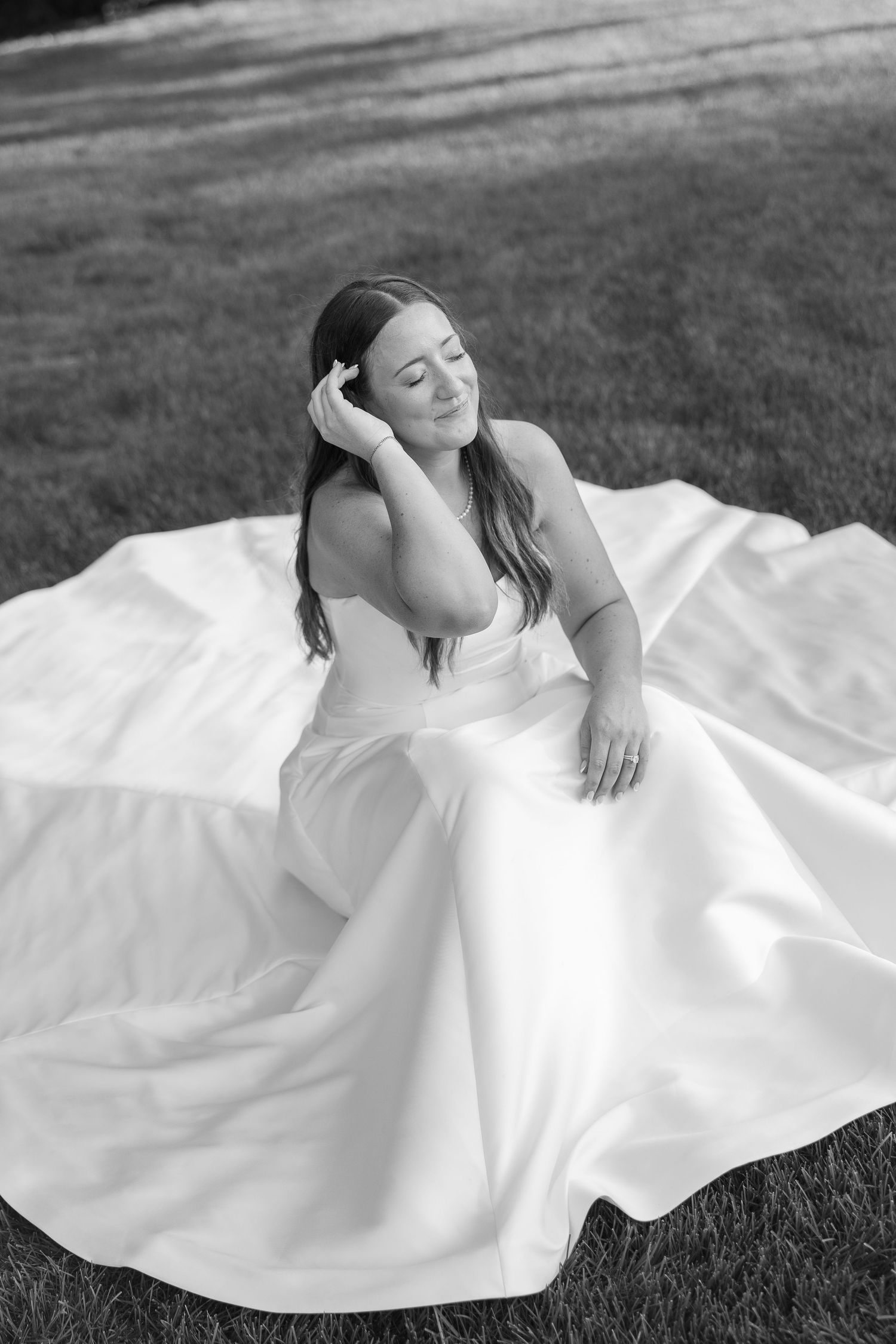 Black and white portrait of bride in flowing white gown sitting gracefully on grass, eyes closed serenely.