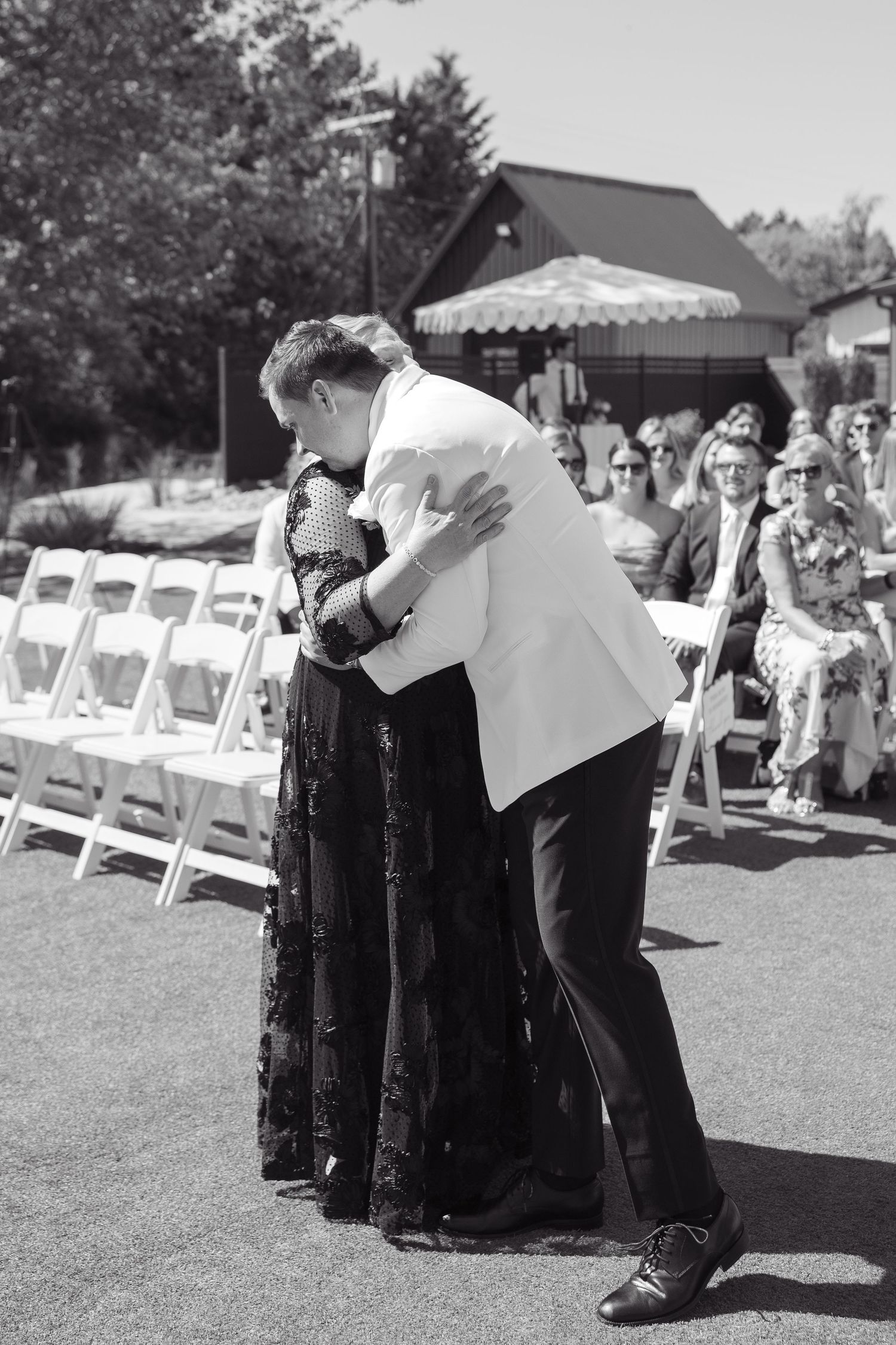 Black and white photo of a couple embracing at an outdoor wedding ceremony with white chairs and guests.