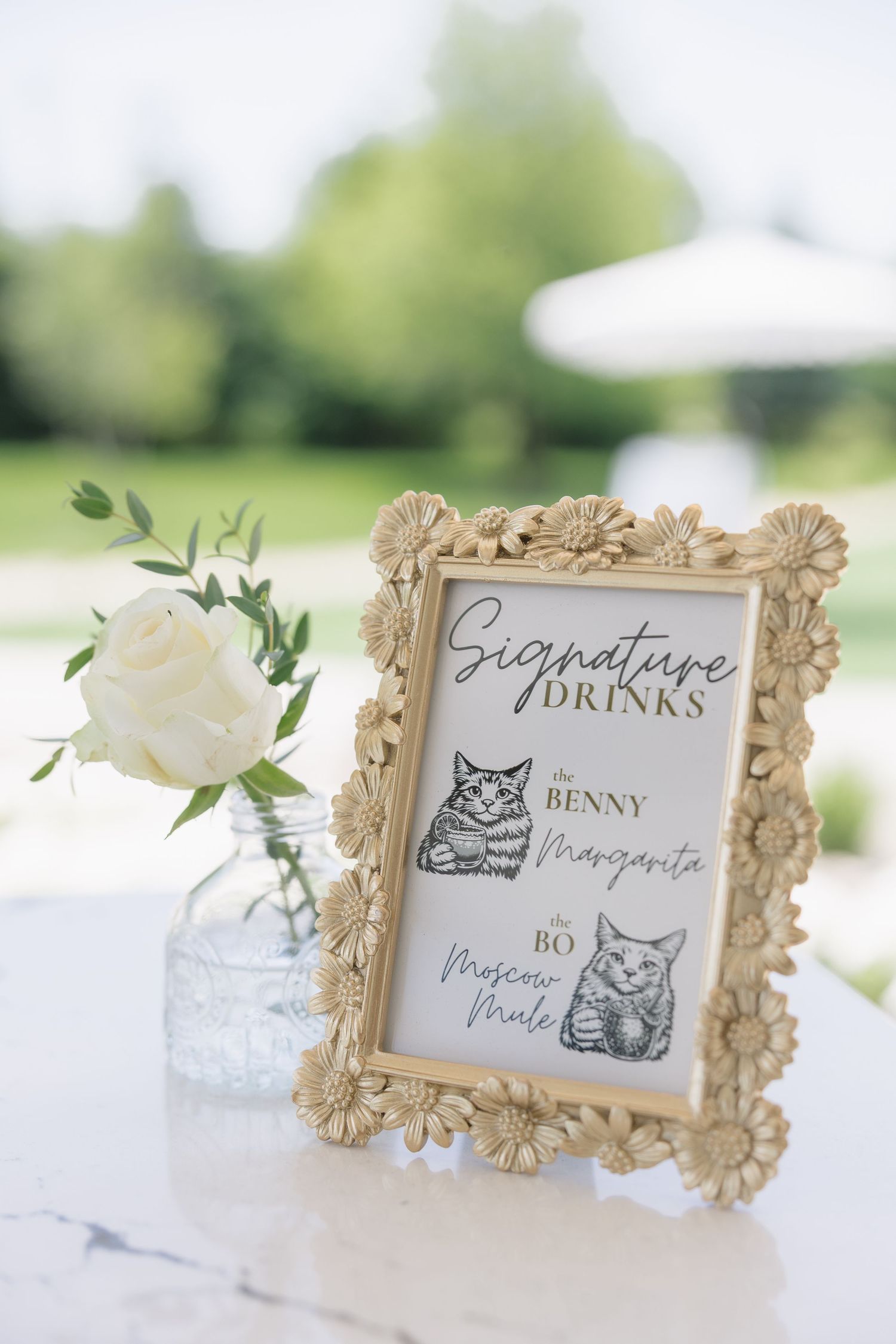 Floral framed signature drinks sign featuring cats named Benny and Bo beside a white rose in a glass vase.
