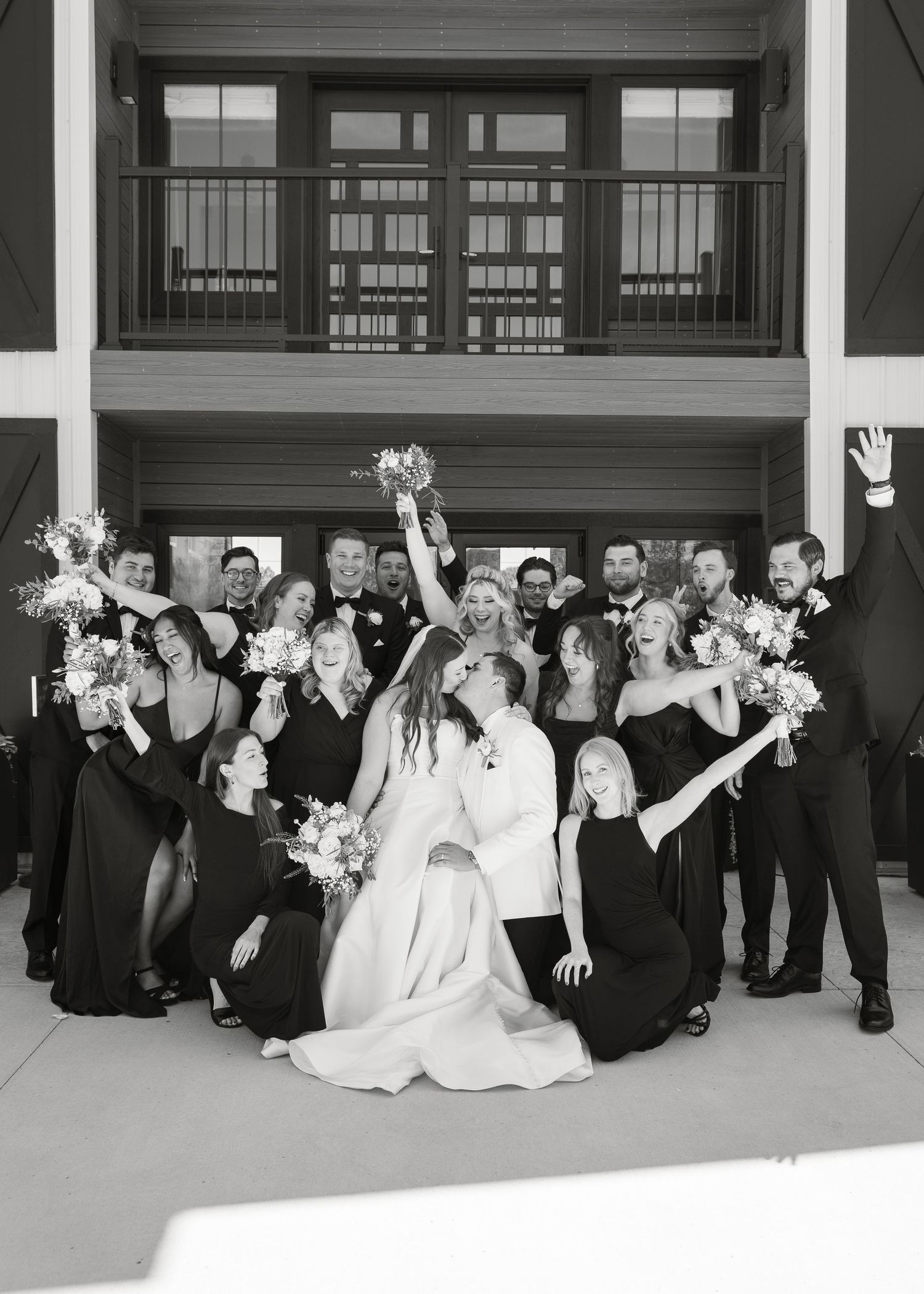 Black and white photo of joyful wedding party celebrating with bride and groom kiss.