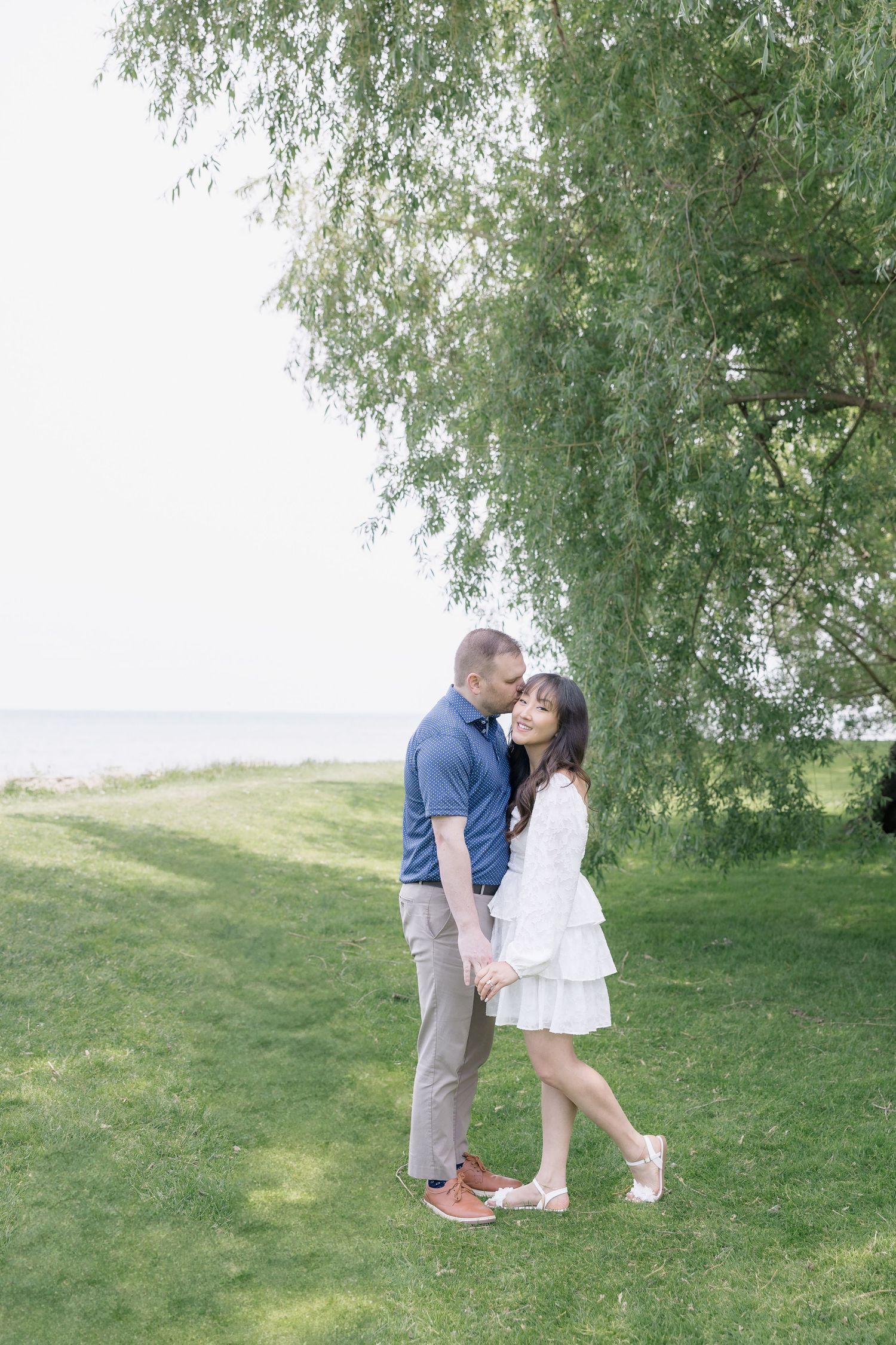 A romantic couple shares playful moments under a weeping willow tree in a grassy park setting.