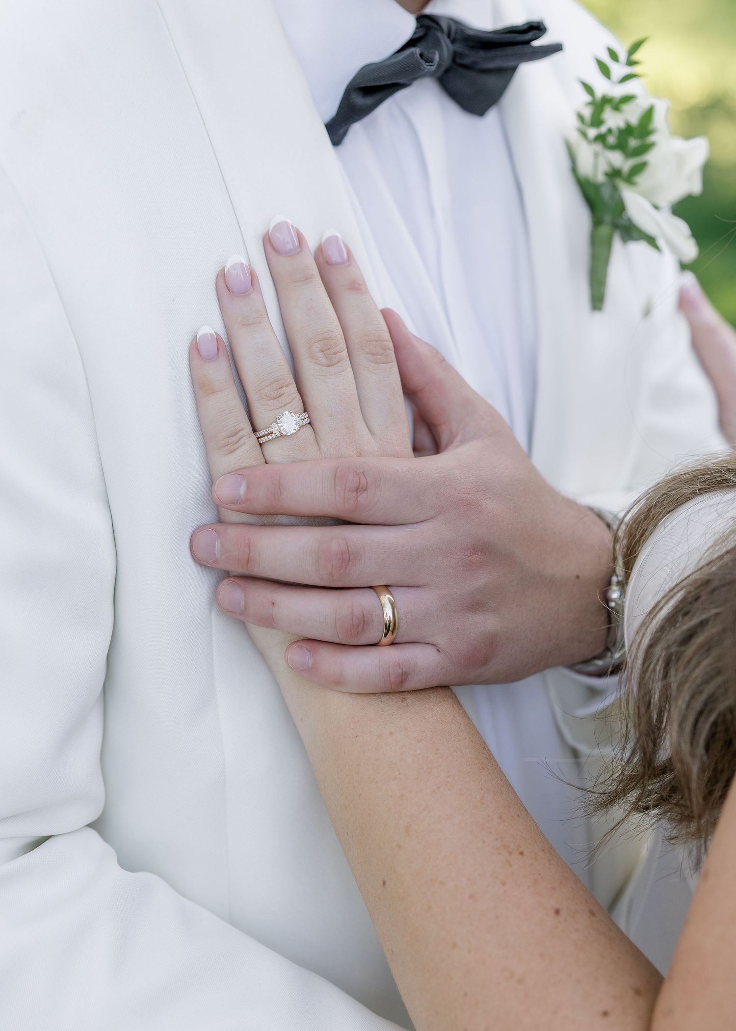 Close-up of newlyweds hands showing wedding rings and engagement ring against the grooms white tuxedo jacket.