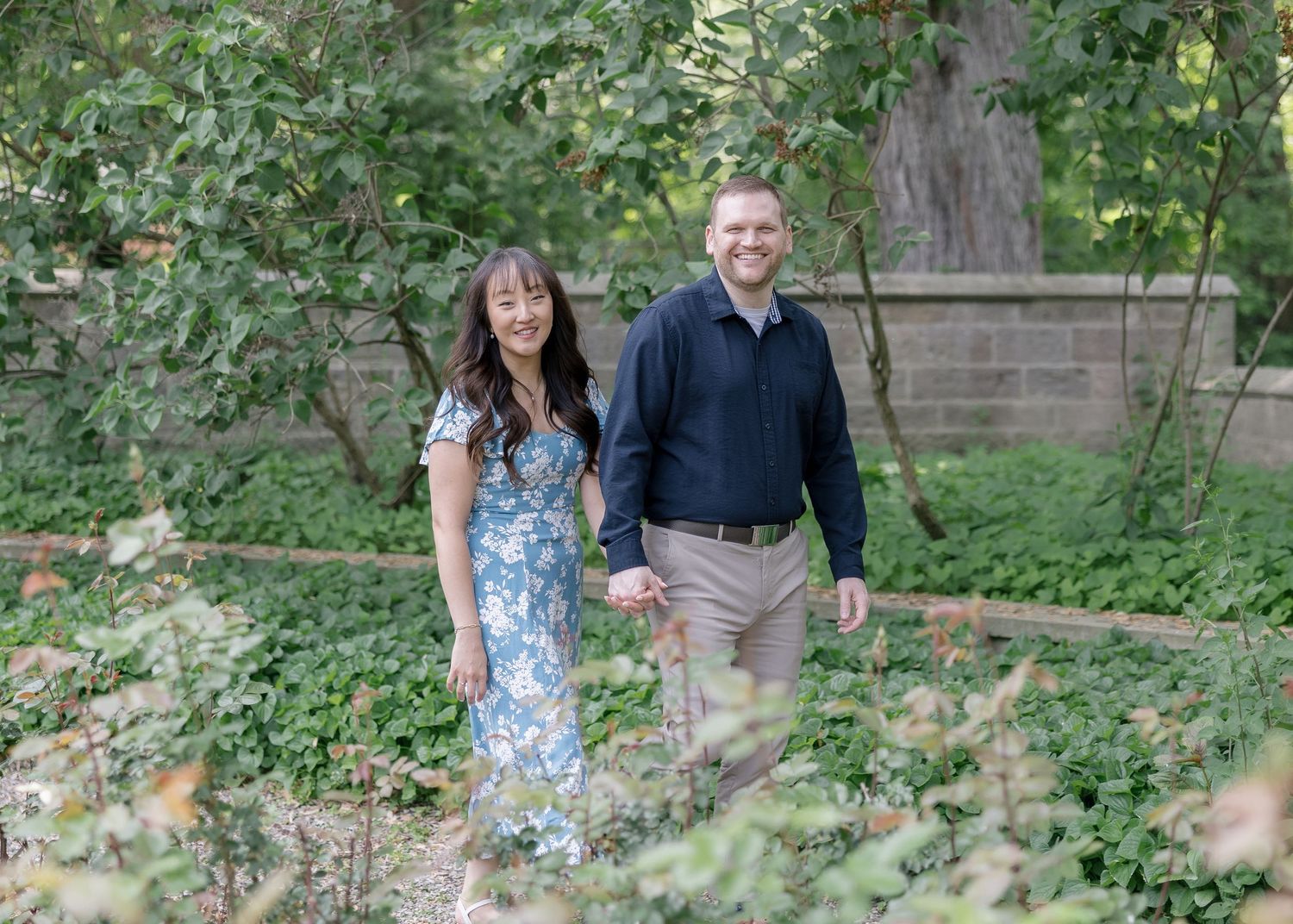 A couple holding hands while standing on a gravel path in a lush garden setting on a sunny day.