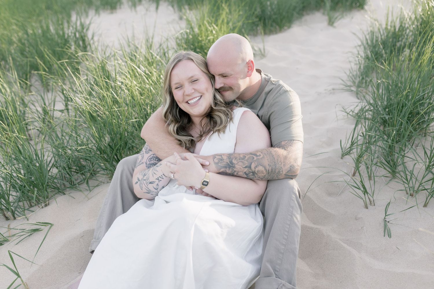 A couple sits together on a sandy beach path surrounded by dune grass in soft dreamy lighting.